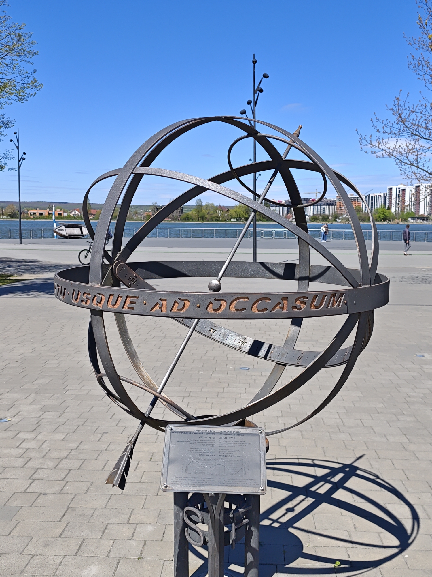 The image depicts an armillary sphere sundial located in Ivano-Frankivsk, Ukraine. The sundial, made of metal, is composed of interlocking rings with the inscription "ORTU USQUE AD OCCASUM" around its equator. A gnomon with an arrow-shaped end extends through the center, casting a shadow on the paved ground. A plaque displays information about the sundial's function and location, providing coordinates of 48°54'46" N and 24°41'37" E.
In the background, a waterfront area is visible, featuring a pedestrian walkway, a bicycle, buildings, and people walking along the embankment. The sky is clear and blue. Trees with budding leaves are present on either side of the frame.