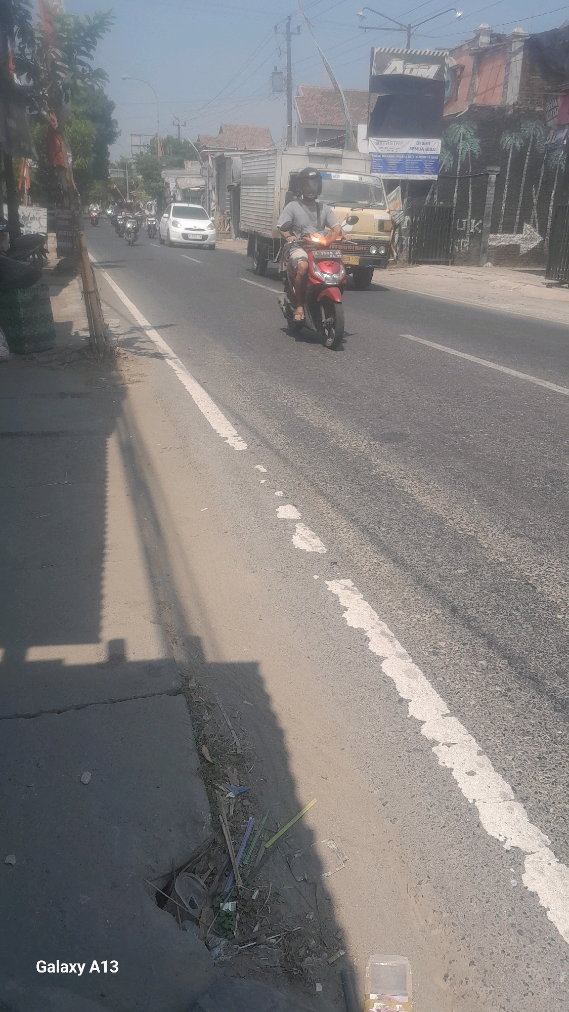A street scene in Mejobo, Indonesia, is depicted on a sunny day. The primary focus is a red scooter carrying a person wearing a helmet and a gray t-shirt. The scooter is on the right side of the road, moving away from the camera. To the left of the scooter, a beige truck is also traveling in the same direction. Further down the road, a white car is visible, and several other motorcycles are in the distance. The road is paved and marked with a white line on the right. The left side of the road features a sidewalk with some vegetation and shadows cast by overhead structures. Some debris, including plastic straws and other refuse, is present on the edge of the sidewalk. Buildings line the right side of the street, including a structure with an "ATM" sign and other commercial establishments. The sky is clear and bright.