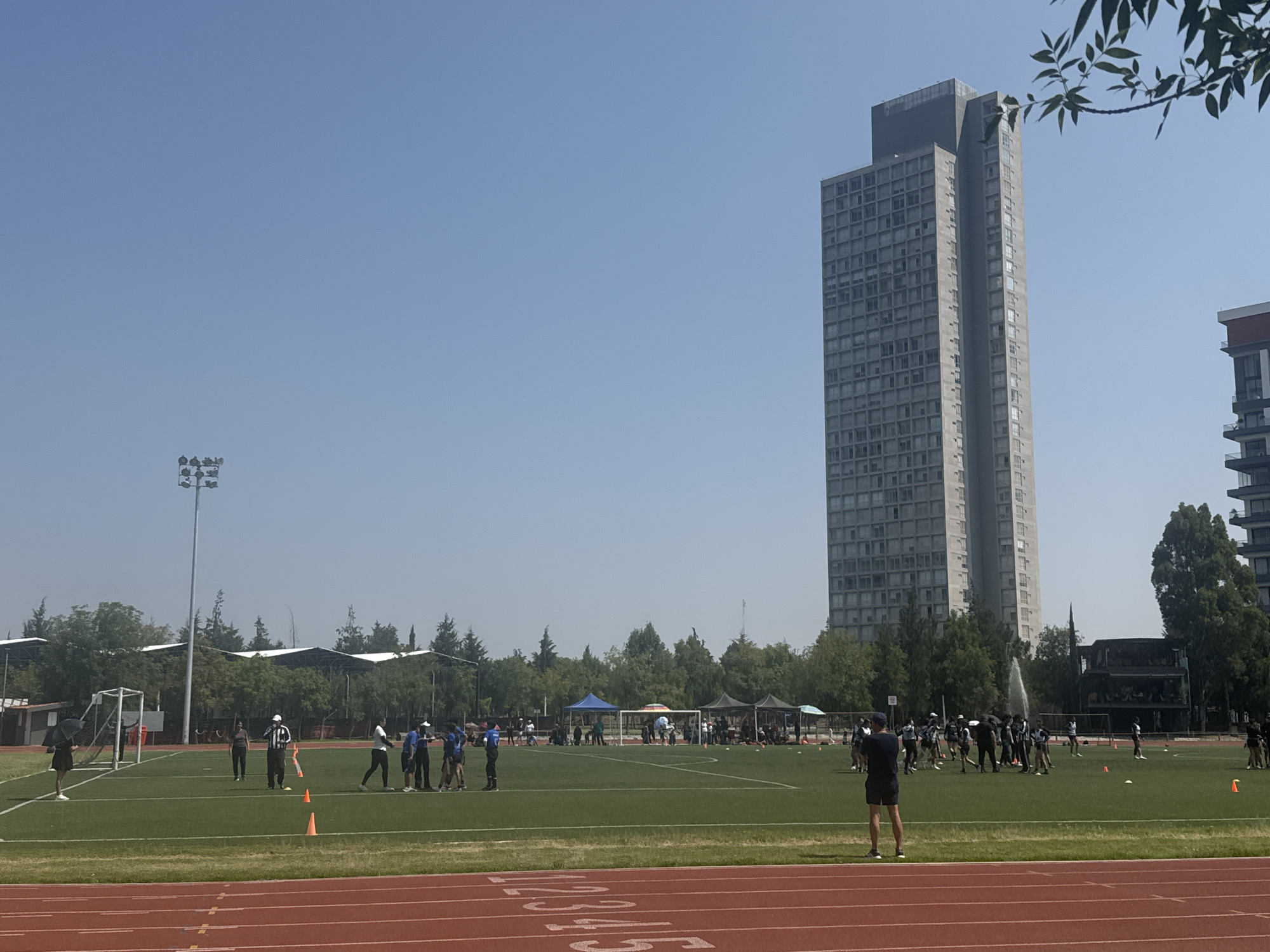 The image shows a sports field in Puebla, Mexico, with a running track in the foreground and a soccer/football field behind. There are several people on the field, some of whom appear to be athletes in uniform and others who are likely coaches or referees. There is a tall, modern-looking building in the background. The sky is clear and blue.