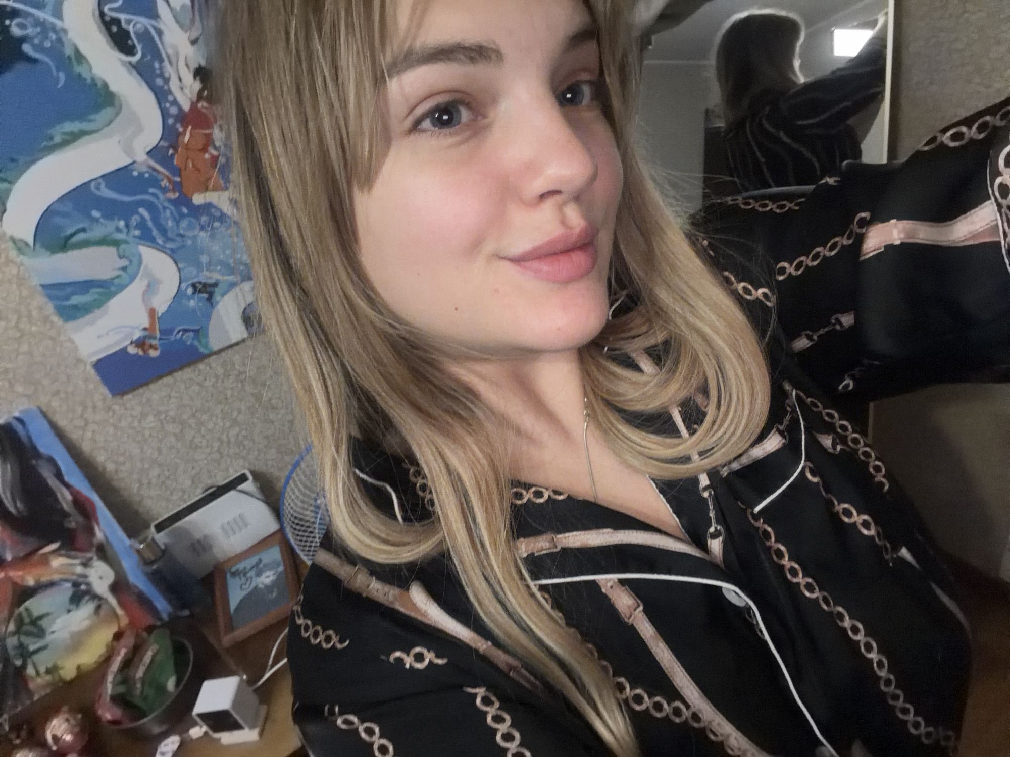 A young woman with blonde hair and bangs is taking a selfie indoors. She is wearing a black button-down shirt with a chain and belt print. In the background, there's a wall with a poster depicting a white dragon and a figure in red, along with other decorative items on a table, including what appears to be a photograph in a frame and a small white box. In the upper right corner, a mirror reflects a silhouette of someone in a similar shirt. The location is likely Naberezhnye Chelny, Russia.