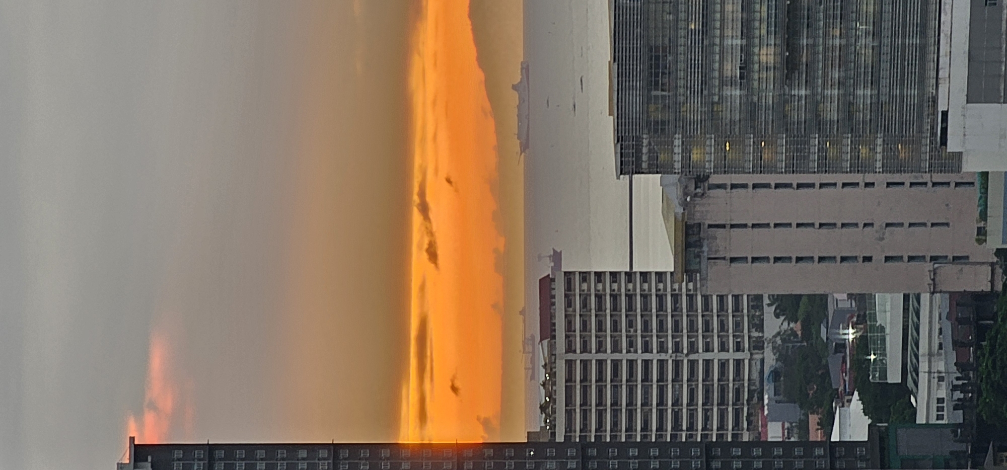 The image shows a cityscape in Makati, Philippines, with a dramatic sunset as the backdrop. The sky is dominated by a vibrant column of orange light, suggesting the sun is either rising or setting behind the clouds. This intense light illuminates the surrounding clouds, creating a surreal and awe-inspiring effect.
In the foreground, the tops of several buildings are visible. The buildings are modern, with distinct architectural features such as rows of windows and what appear to be balconies or external structural elements. The lighting from the sunset casts a warm glow on the buildings, highlighting their textures and forms. The lower portion of the image reveals the density of the urban environment, with multiple structures packed closely together.
The time of day is clearly evening or early morning, indicated by the sunset's colors. The weather appears to be partly cloudy, with the clouds acting as a canvas for the dramatic sky phenomena. The location is identified as Makati, Philippines, a major business and financial hub. While no people are explicitly visible, the presence of numerous office buildings and residential structures implies a populated urban area. The overall mood is one of natural beauty contrasting with the man-made urban landscape, evoking a sense of wonder and the power of nature. No visible text is present in the image.