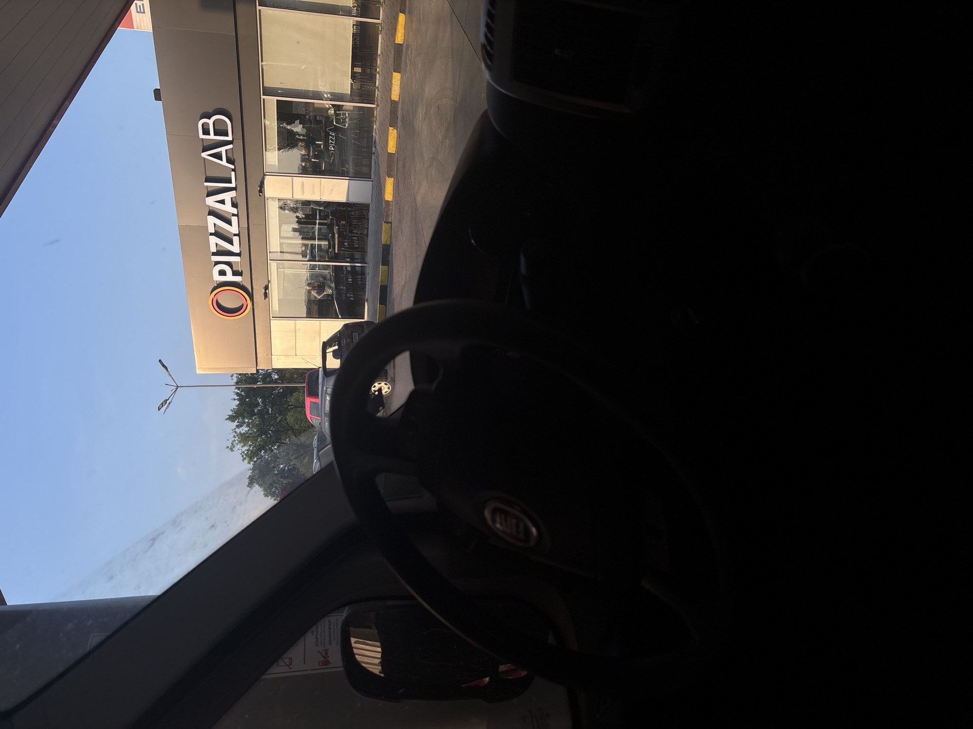 The image provides a driver's-eye view from inside a vehicle, looking towards a commercial establishment in Tsaratsovo, Bulgaria, during what appears to be late afternoon or early evening under a clear blue sky.
The immediate foreground, occupying the lower half of the frame, is dark and shows the interior of the vehicle. A dark, three-spoke steering wheel with a partially obscured central logo (consistent with a Fiat emblem) is prominent on the right. To the left, a dark side mirror and the vehicle's A-pillar are visible. A white sign, possibly a warning sticker inside the vehicle or at a gas pump, shows text including "СТАРТИРАЙ" (Start) and "DO NOT START" in dark letters.
Beyond the vehicle, a paved service area or parking lot stretches towards a single-story commercial building. The paved surface features yellow and black curb blocks separating the driving area from the building's front. Several vehicles are parked in the distance, including a dark hatchback on the far left, a red SUV, and a dark sedan. A streetlight with two luminaires stands tall against the sky on the left, with green trees visible behind it and the parked cars.
The central focus of the background is a modern building with a dark grey upper facade and large, floor-to-ceiling glass windows below. A prominent sign on the facade reads "O PIZZALAB" in white capital letters, with the 'O' stylized as an orange-rimmed circle. Through the glass windows, the interior of the Pizzalab restaurant is visible. Dark tables and chairs are arranged inside, and several individuals are present, appearing to be patrons or staff. One person with dark hair is distinguishable near a counter on the left side of the interior. A small interior sign with "PIZZA" is also visible. To the far right, above the Pizzalab sign, a portion of a red and white canopy or structure with a visible "E" (possibly part of "EL" for a fuel brand) indicates the presence of a gas station adjacent to or co-located with the restaurant.