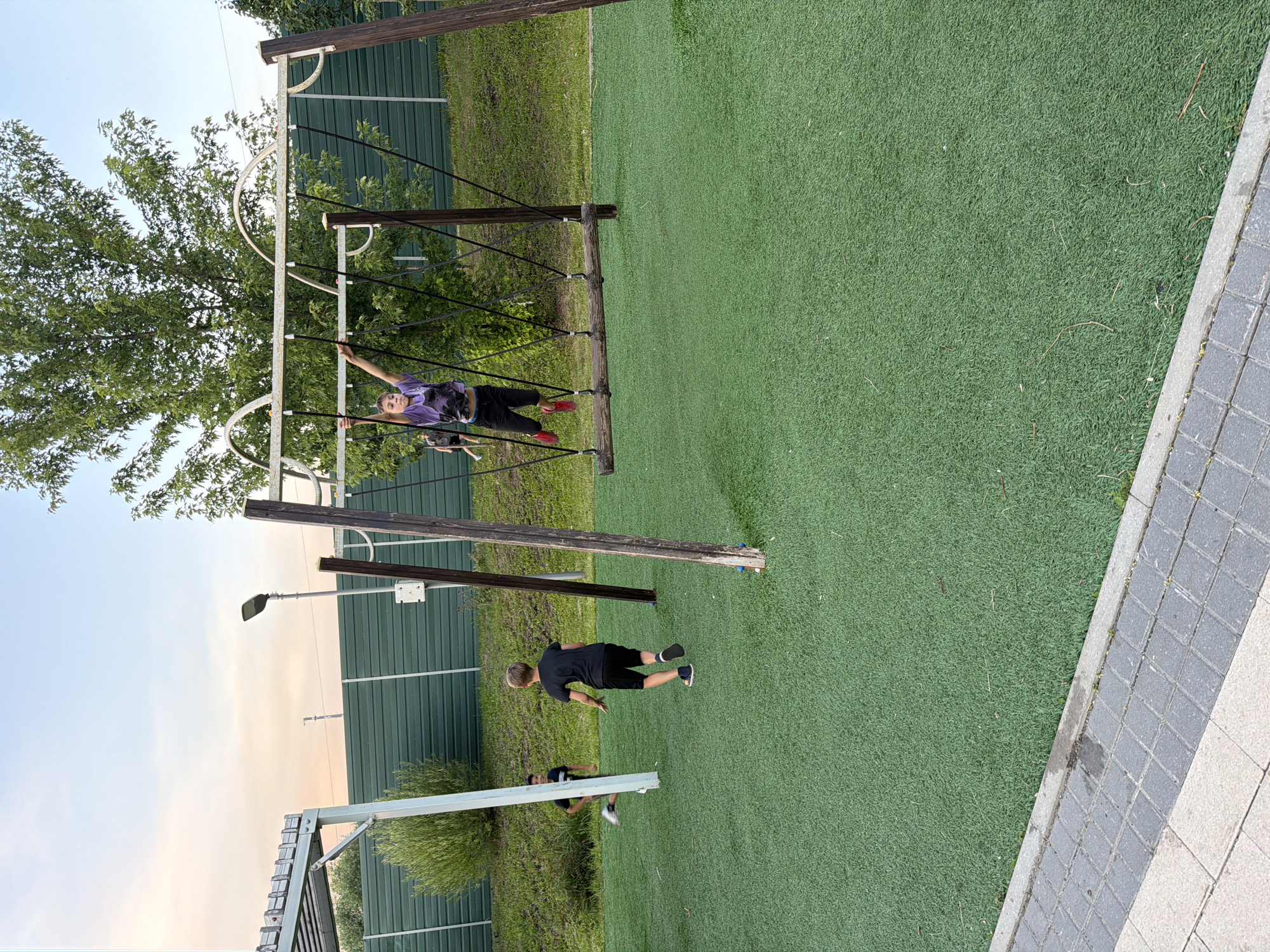 A boy in a purple shirt and black pants is swinging on a wooden swing set in a park. He is in mid-air with his legs extended. Two other boys are on the ground in the background, one running and the other standing. The ground is covered in green artificial turf. Behind the swing set is a green metal fence and trees. The sky is light blue with some clouds. The scene appears to be in a park in the city of Shcherbinka, Russia.