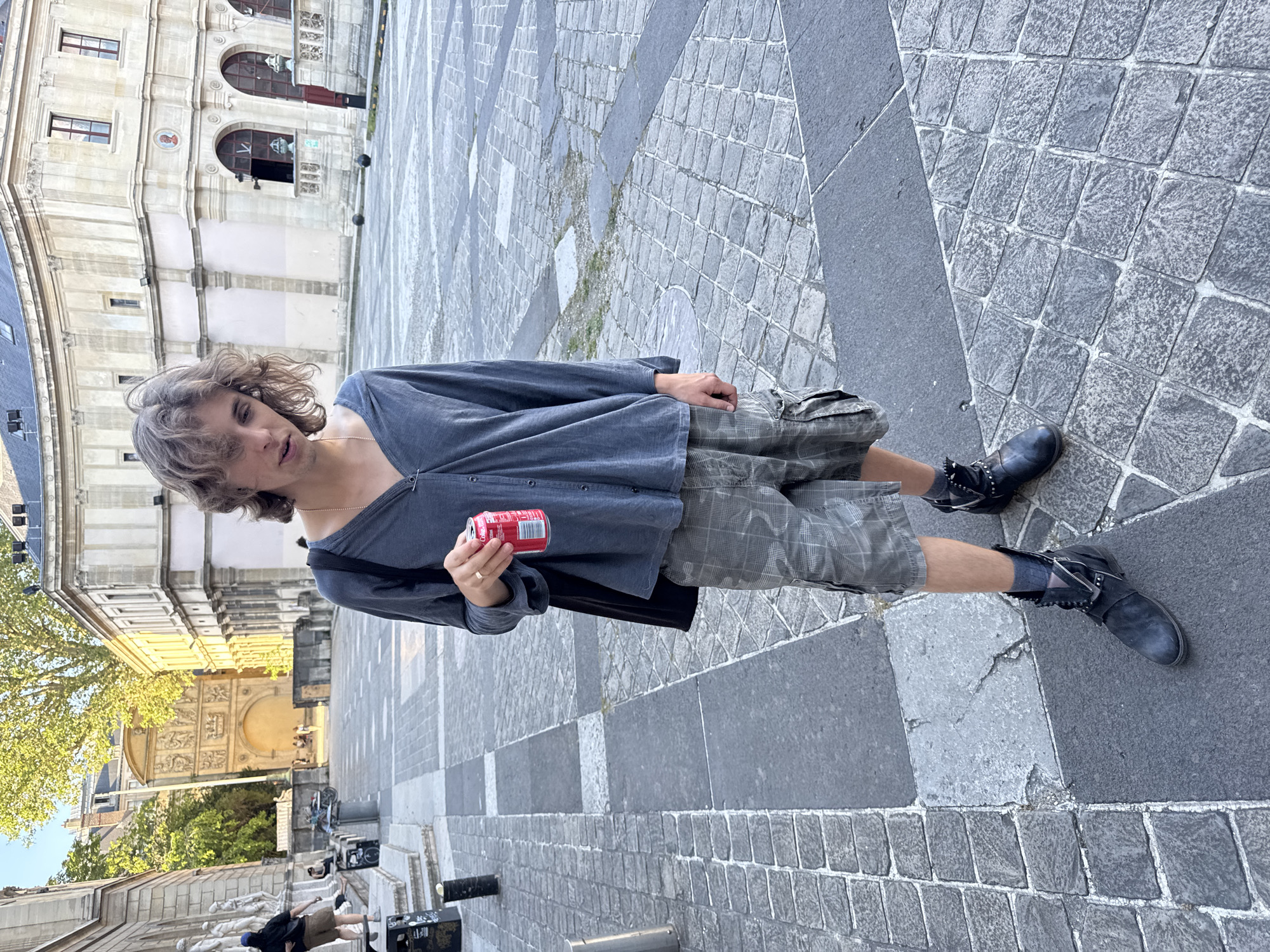 The image shows a person standing on a stone-paved surface, likely a plaza or square. The person is holding a red can, possibly a soda or energy drink. The person wears a light grey shirt and checkered shorts with black boots. The person stands in front of a large, ornate building typical of Parisian architecture, suggesting the photograph was taken in Paris, France. The background also includes trees and other buildings.
