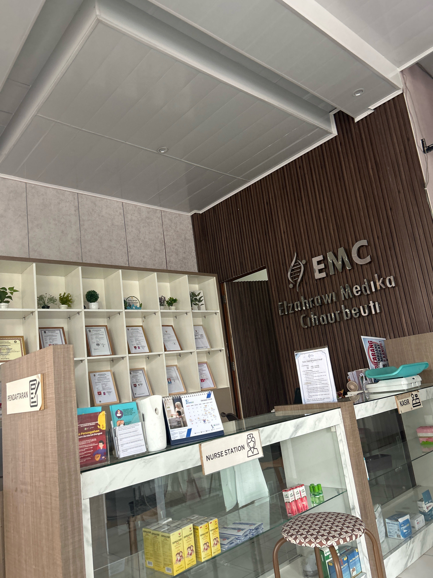 The image depicts the interior of a medical facility, likely a clinic or hospital reception area, in Sukasetia, Indonesia. The overall impression is one of clean, modern design with a focus on organization and information display.
Dominating the background is a wall with vertical wooden paneling, upon which a prominent metallic sign reads "EMC Elzahrawi Medika Cihauabeuti." This sign indicates the name and location of the medical institution. To the left of the wooden wall, a white shelf unit is visible, filled with numerous framed certificates and diplomas. Scattered amongst the certificates are small decorative potted plants.
In the foreground, a reception counter stretches across the width of the image. On the left side of the counter, a wooden panel features a sign with the word "PENDAFTARAN" (Registration) accompanied by an icon of a clipboard and pen. Beneath this sign, various informational brochures are displayed. The counter itself has a marbled white surface.
To the right of the registration area, a glass display case showcases medical supplies and products, including boxes of medication and what appear to be baby scales. A small stool with a patterned cushion sits in front of the counter. Further to the right, another sign indicates "NURSE STATION" with a corresponding icon.
The ceiling is a white, paneled structure with recessed lighting. The environment appears to be well-lit and professional. No people are visible in the image. The scene captures a functional and welcoming reception area of a medical establishment.