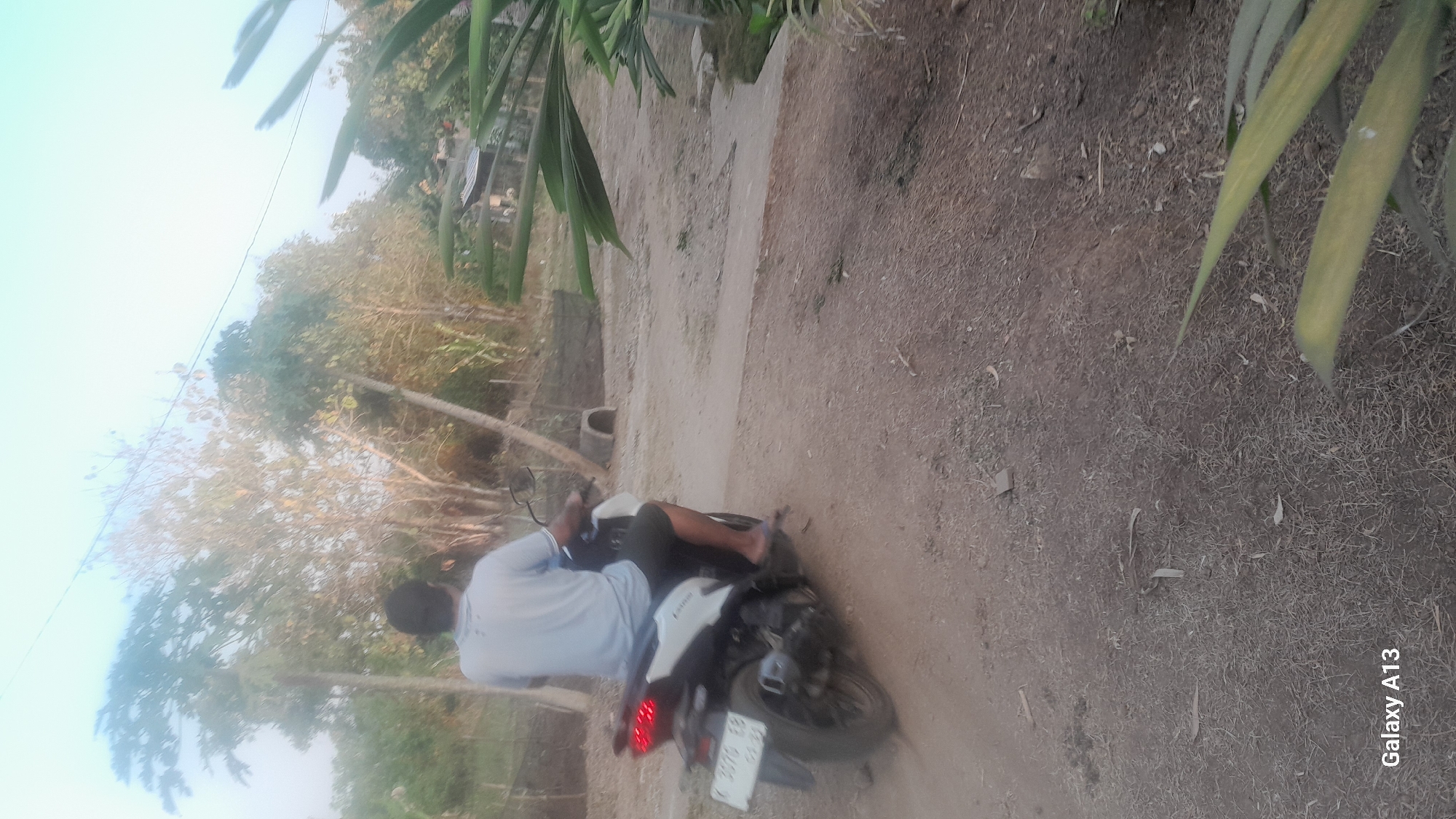 A person is riding a white and black scooter on a dirt road. The person is wearing a light blue t-shirt and dark shorts. The scooter has a license plate that reads "K 3076 EB". The road is surrounded by trees and vegetation, with a light sky in the background. There is a small concrete structure visible to the left of the road. The scene appears to be in a rural or semi-rural area. The location is Hadipolo, Indonesia.