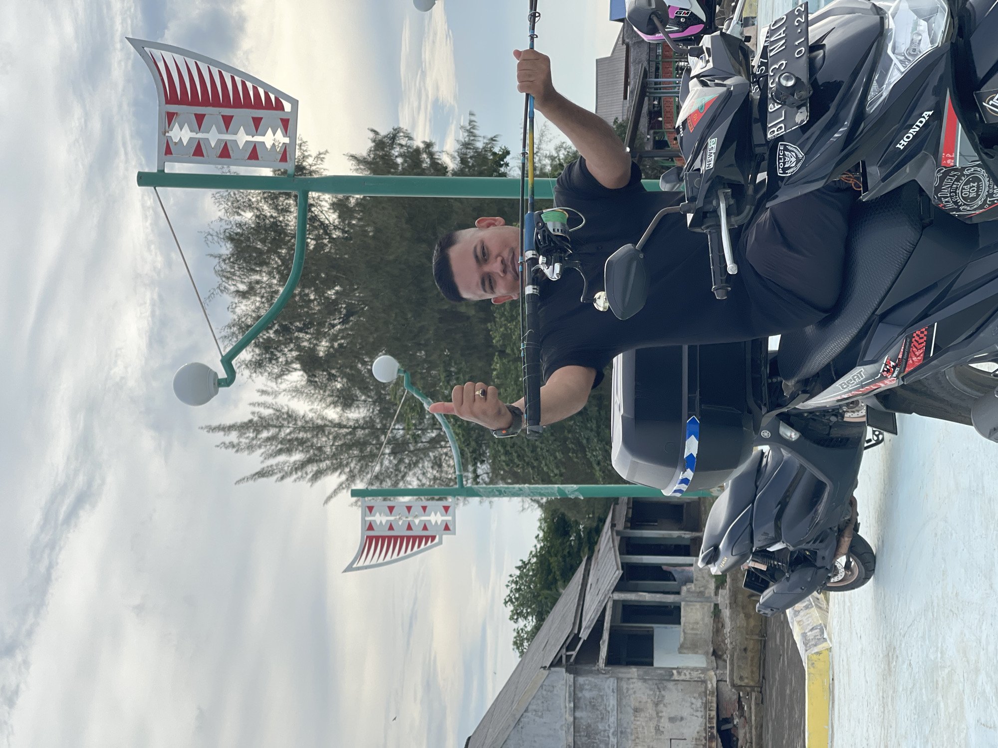 This is a portrait-oriented, slightly tilted photo of a man holding a fishing rod. He is sitting on a black motorcycle, which is parked next to a light-colored wall. The man is wearing a black t-shirt and a watch. He is smiling and giving a thumbs-up gesture. The motorcycle has various stickers and a license plate that reads "BL3 NAC 01-22". In the background, there are trees and a cloudy sky. Two decorative flags with a red and white sawtooth pattern are visible, mounted on green poles. A building with a weathered appearance is also in the background. The overall scene suggests a casual outing, possibly near a coastal area given the general ambiance and the fishing rod. The time of day appears to be daytime, with diffused light from the overcast sky. The location is identified as Lancok, Indonesia.