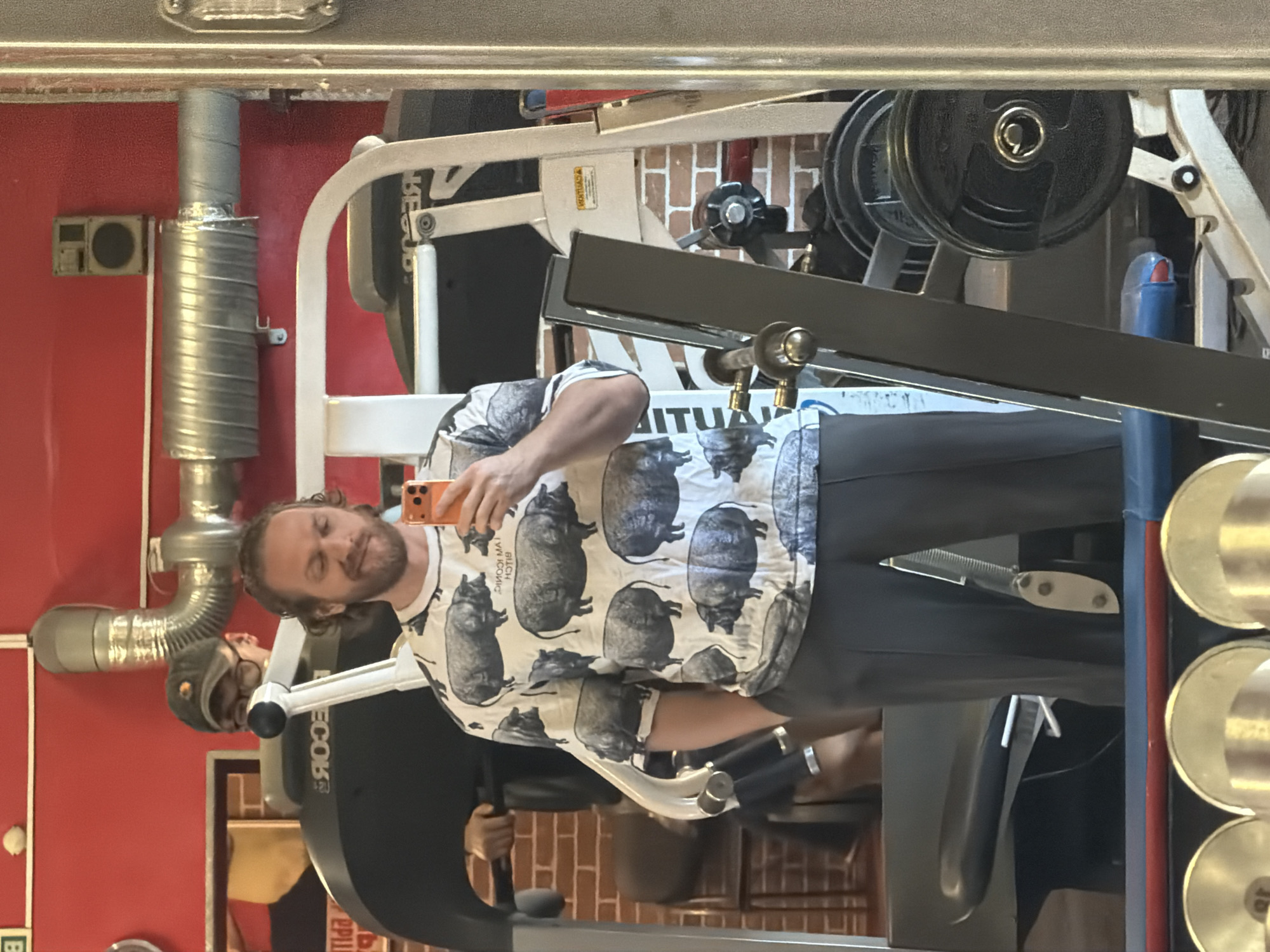 A man with long reddish-brown hair and a beard is taking a selfie in what appears to be a gym. He is wearing a t-shirt with images of pigs printed on it and dark pants. He is holding his orange phone in his right hand. Behind him is gym equipment, including weights and a treadmill, and a brick wall. The red wall behind the equipment suggests the scene is likely in a gym or exercise facility. Due to lack of clear indicators, the exact location in Saint Petersburg, Russia cannot be precisely determined.