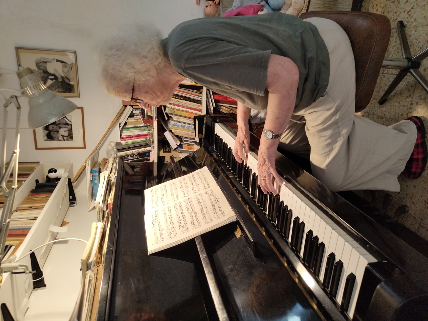 An elderly woman with white, curly hair and glasses is playing a piano in a room filled with books. She is wearing a green t-shirt and light-colored pants, and her feet are clad in red and black plaid slippers. Her hands are on the piano keys, and there is a sheet of music in front of her on a stand. The piano is black, and the keys are a mix of white and black. The room has white walls and is lined with bookshelves filled with books. There are framed photographs on the wall, one showing two young children, and another of an adult and a child. A desk lamp is attached to a shelf above the piano. In the background, there are some dolls visible. The overall impression is one of a cozy, lived-in space dedicated to music and literature. The scene is likely a home in Jerusalem, Israel, given the presence of bookshelves, a piano, and the general domestic setting.