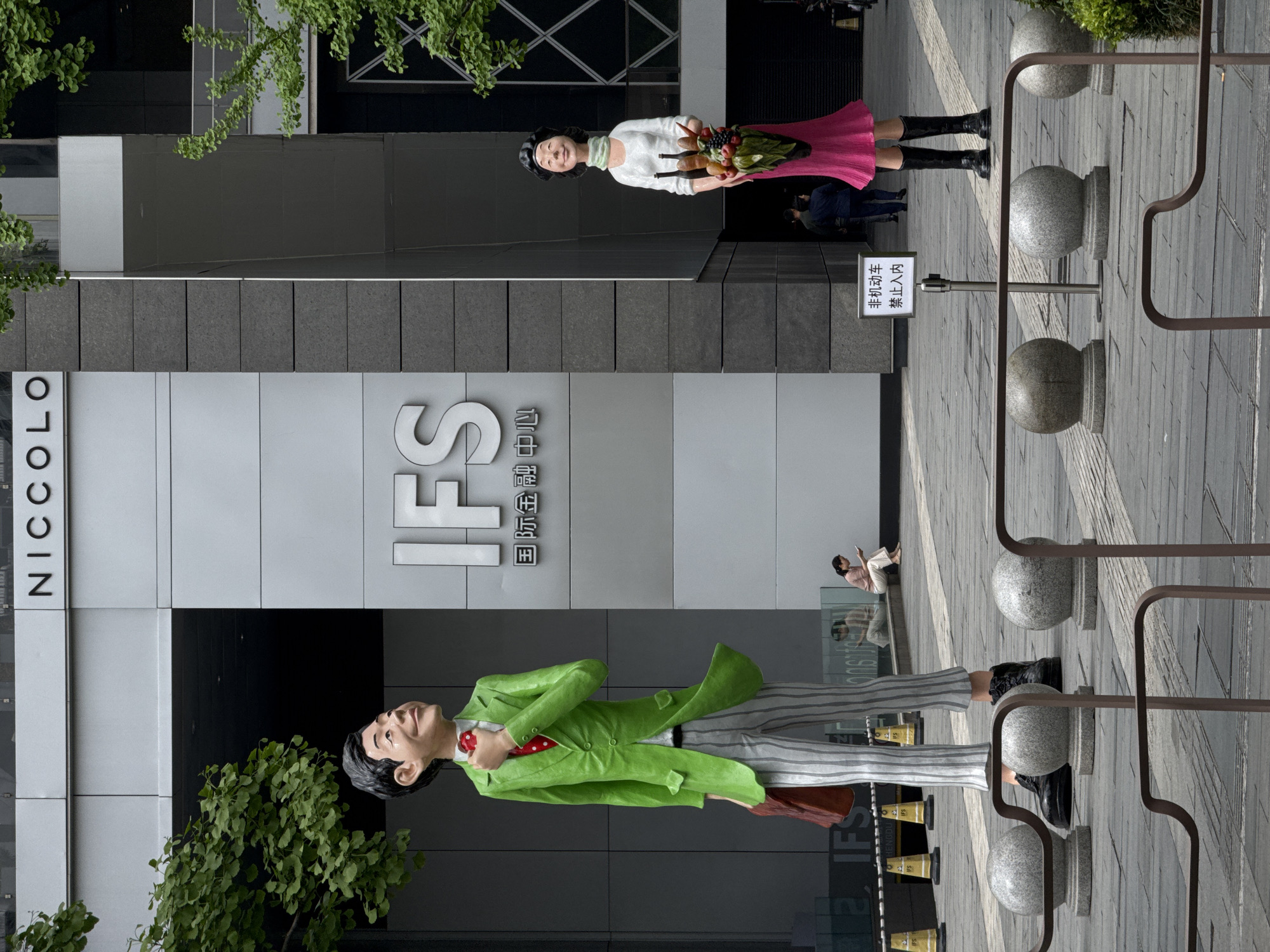 The image shows the IFS (International Finance Square) shopping mall in Jinjiang District, Chengdu, China. The building facade features the "NICCOLO" name vertically on one section and the "IFS" logo on a larger, grey section. Two large cartoon statues stand in front of the building: a man wearing a bright green suit and a woman in a pink skirt holding a fruit basket. A sign with Chinese characters is visible near the statues. Pedestrians are present in the background. The ground is paved with stone tiles, and there are decorative stone sphere posts with metal railings. Trees are positioned on either side of the building.