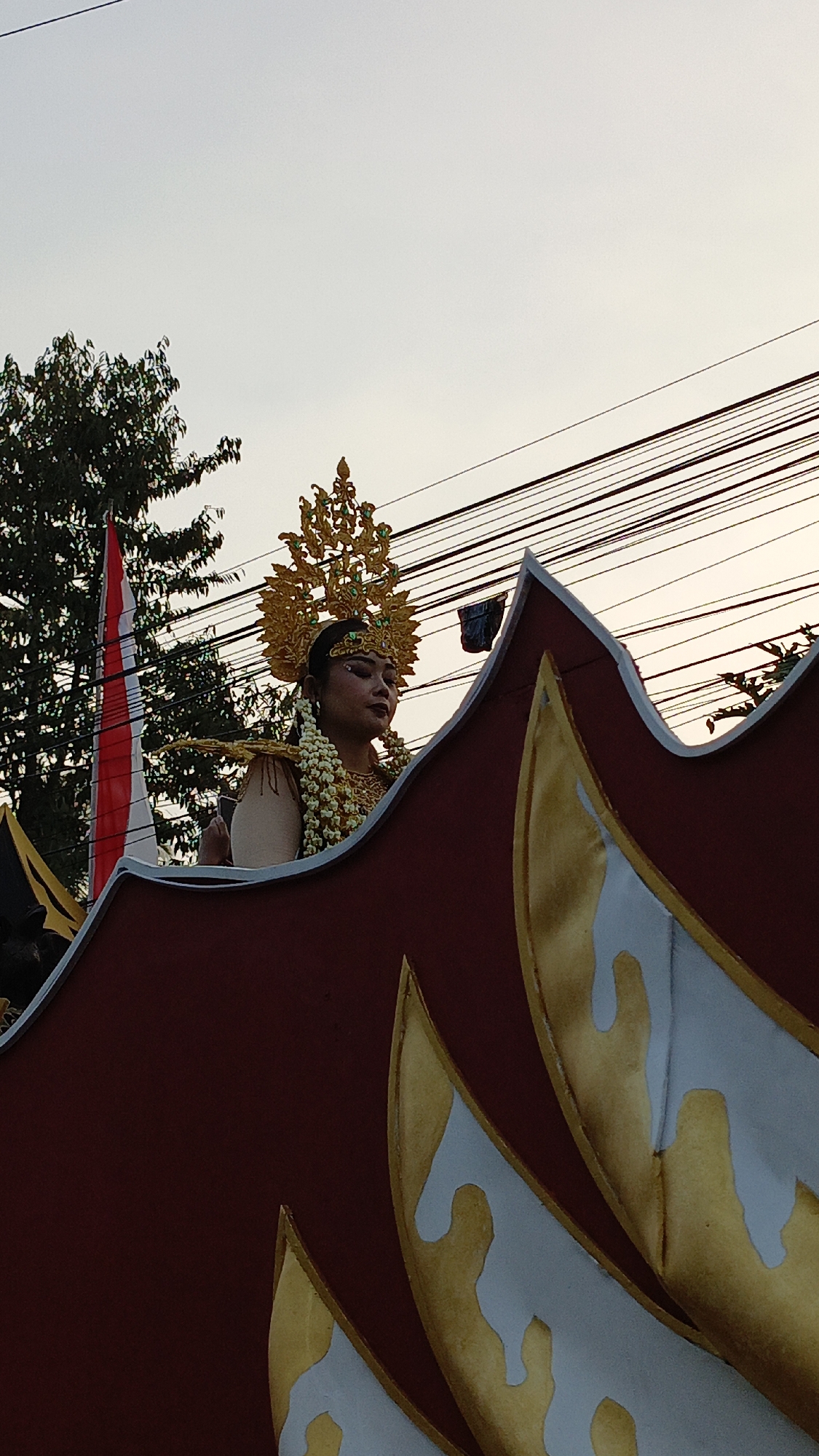 A young woman with a golden headdress adorned with green stones and a floral garland around her neck is positioned on a float decorated with red and gold motifs. She wears traditional makeup and a light-colored garment. To her left, a red and white Indonesian flag is visible, with a tree in the background. Overhead, a dense network of electrical wires stretches across the sky. The float appears to be part of a parade or festival in Krajan Lor, Indonesia, given the context of the image.