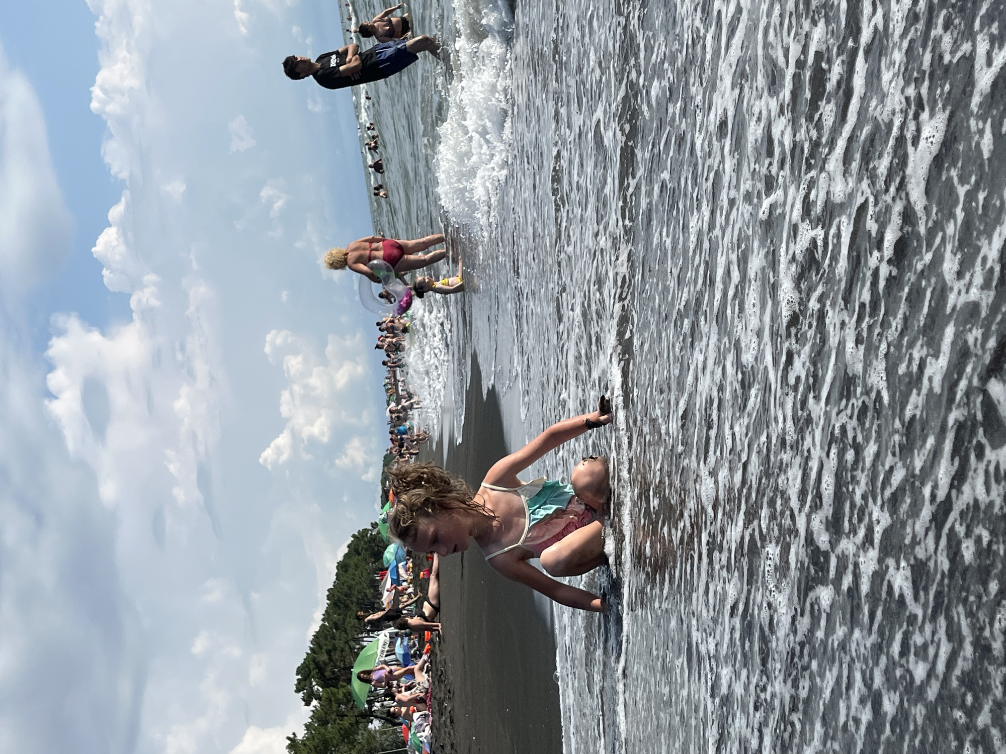 In Ureki, Georgia, a young girl with curly blonde hair sits in the shallow water of a crowded beach, facing right and looking out towards the sea. She wears a light-colored swimsuit with pastel stripes. The sea is calm, with small waves lapping at her. Behind her, numerous people are scattered across the dark sand and in the water, many under colorful umbrellas. Trees line the edge of the beach in the background, under a cloudy sky.