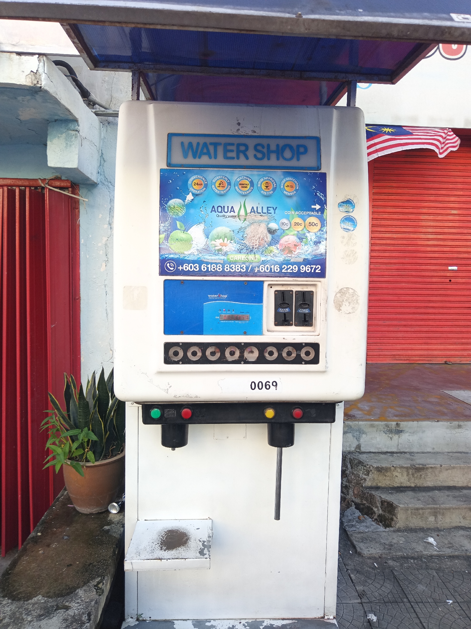 A white water vending machine, labeled "WATER SHOP," stands outdoors. It is located in Kuala Lumpur, Malaysia. The machine features a blue display with text such as "AQUA ALLEY" and a phone number, and coin slots for 10c, 20c, and 50c. Below the display are five circular dispensing nozzles and a serial number "0069". A small ledge is positioned beneath the nozzles. To the left of the machine, a potted snake plant is visible on a concrete ledge. The background includes a red corrugated metal gate, a building with blue and white paint, and concrete steps. A Malaysian flag is visible on the right side of the building.