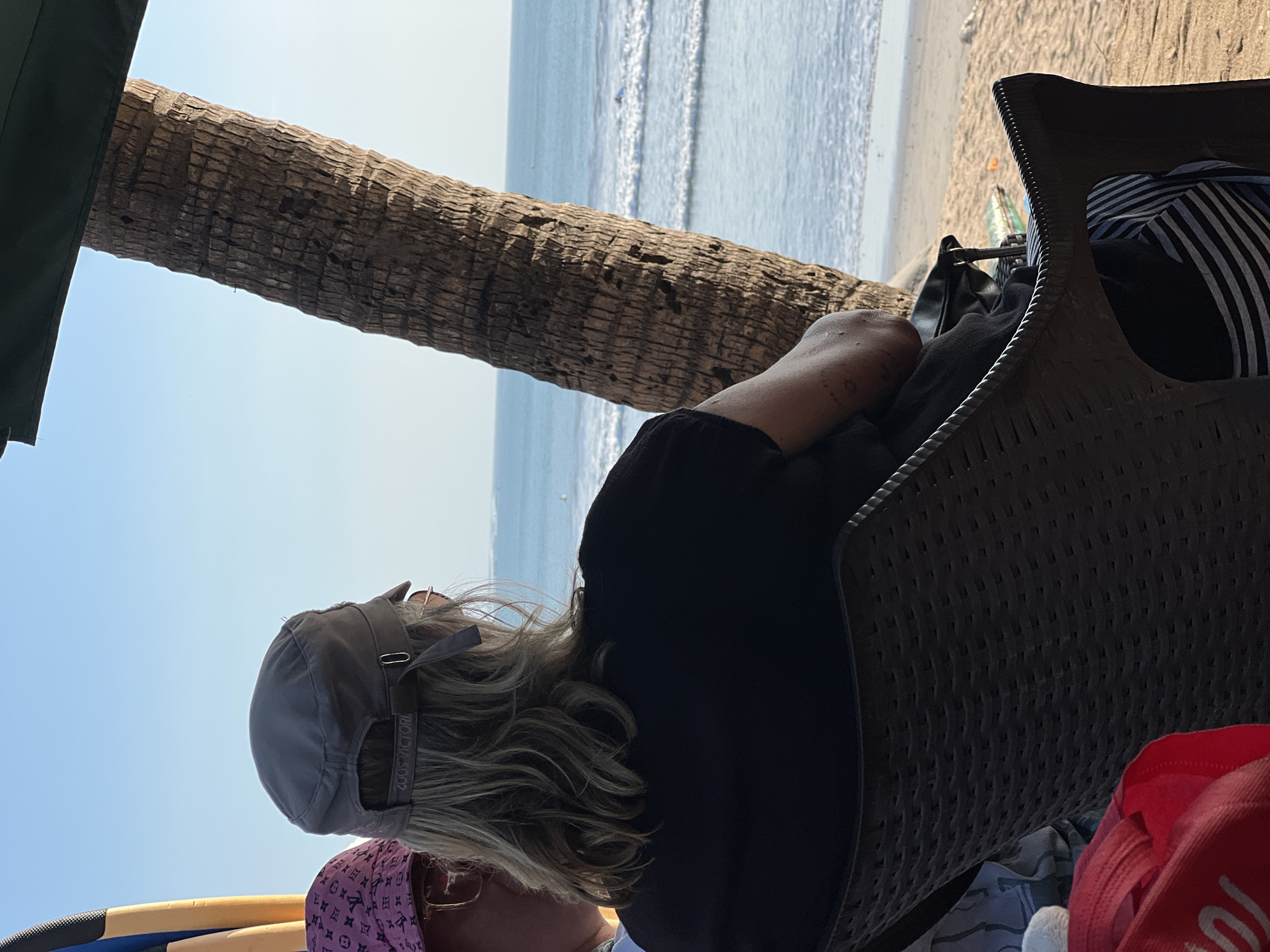 Two individuals are seated on chairs on a sandy beach, with the ocean and horizon visible in the background. The person in the foreground, facing away from the camera, wears a black t-shirt, a grey baseball cap with "ZOOS VICTORI" visible on the strap, and striped pants. Their arm has visible tattoos. To their left, partially visible, is another person wearing a pink hat with a Louis Vuitton pattern and eyeglasses. A surfboard with blue and yellow accents stands upright to the left of the individuals. A palm tree trunk occupies the right side of the frame. The scene appears to be a relaxed setting, possibly a beachside cafe or seating area in Kuta, Indonesia, given the presence of a surfboard and tropical elements.