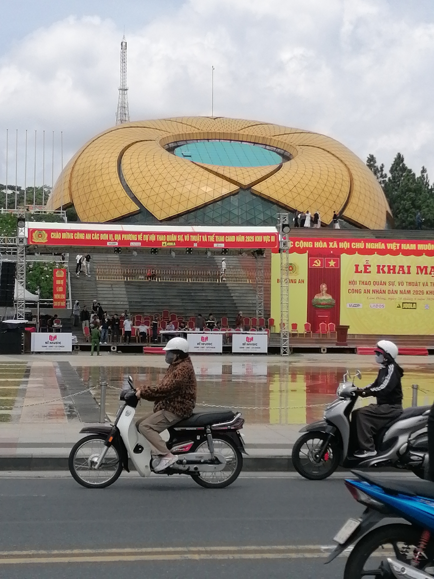 A wide-angle, eye-level shot captures a busy outdoor event in Da Lat, Vietnam, with a distinctive lotus-shaped building as the backdrop. In the foreground, two individuals on motorbikes navigate a wet, reflective street. The person on the left rides a dark and white motorbike, wearing a leopard-print jacket and a white helmet. To their right, another person on a silver motorbike is also wearing a helmet and dark clothing. The street appears recently rained on, with puddles reflecting the surrounding environment.
Behind the motorbikes, a large, stepped seating area is visible, populated by a crowd of people. Red chairs are neatly arranged on the steps. At the top of the steps, a large banner with Vietnamese text and a golden lotus design is prominently displayed. To the right of this banner, a larger yellow banner features an image of Ho Chi Minh and more Vietnamese text. A stage-like structure with scaffolding and speakers is set up on the left side of the scene.
The iconic lotus-shaped building dominates the background, its golden exterior panels forming intricate geometric patterns. A turquoise central dome is visible within the opening of the lotus. A tall, slender tower, likely a radio or communication tower, rises above the building against a cloudy sky. Green trees are visible in the distance, framing the scene. The overall impression is of a vibrant public gathering taking place in a city known for its unique architecture and pleasant climate.