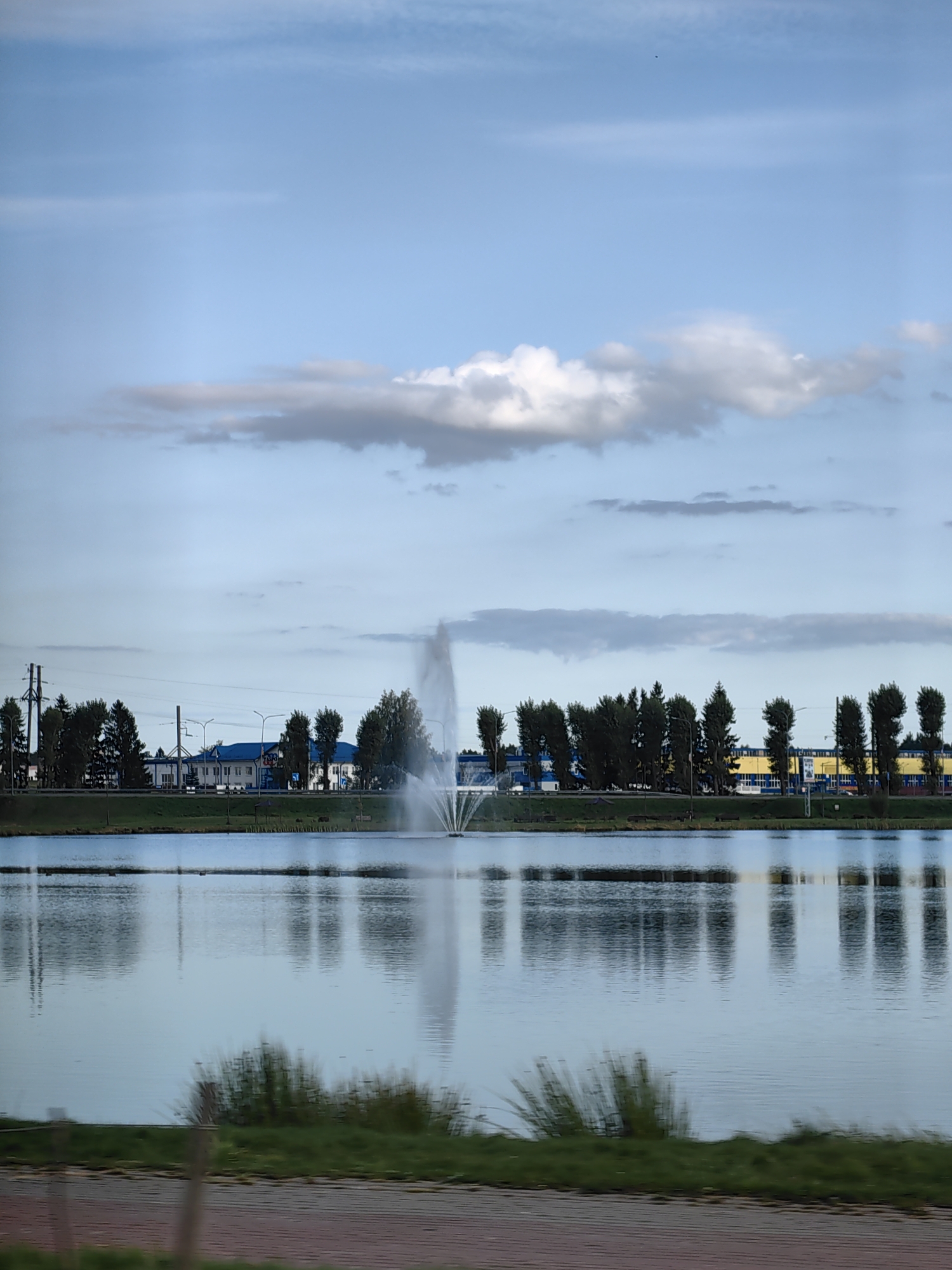 A wide shot depicts a tranquil body of water in Dzyarzhynsk, Belarus, reflecting a cloudy sky and the adjacent cityscape. The water's surface is calm, disturbed only by a tall, white fountain erupting in the middle distance, its spray catching the light. Along the far bank, a line of trees stands tall, interspersed with various buildings. One prominent building features a bright blue roof, while others are a mix of white and yellow. Utility poles and wires are visible, indicating the presence of infrastructure. The foreground shows a glimpse of a paved path and green grass, suggesting a park or public area. The overall scene is one of urban tranquility, with nature and man-made structures coexisting by the water's edge.