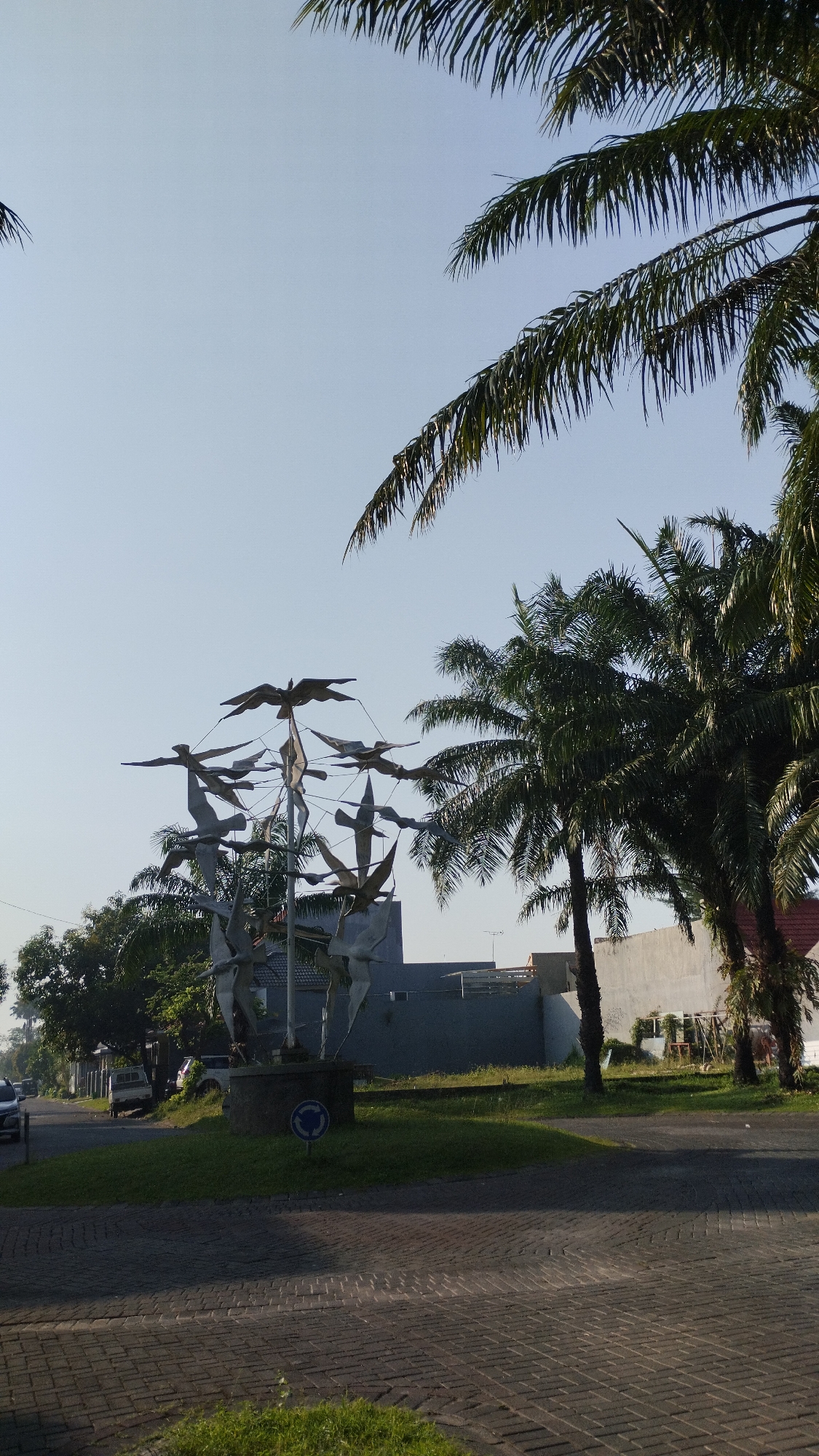 The image depicts a street scene in Sidodadi, Indonesia, bathed in bright, clear daylight under a pale blue sky. In the foreground, a textured brick-paved road with scattered patches of green grass occupies the lower portion of the frame. To the left, a circular grass island is situated, featuring a modern sculpture composed of multiple stylized bird figures arranged in a circular, upward-sweeping formation. A small blue traffic sign indicating a roundabout is visible on this island. Beyond the island, a two-lane asphalt road curves to the left, with a few parked vehicles, including a white truck and a white car, visible on the side. Lush green trees line the left side of the road.
The midground is dominated by a long, light-colored concrete wall that stretches horizontally across the frame. Behind the wall, several palm trees with their characteristic fronds are prominent. The largest palm trees are on the right side of the image, with their dark green foliage contrasting against the sky. More trees and some buildings are visible behind the central sculpture and to the left. The sky is a uniform, bright blue, with no clouds apparent. The overall impression is a bright, sunny day in a tropical or subtropical urban environment.