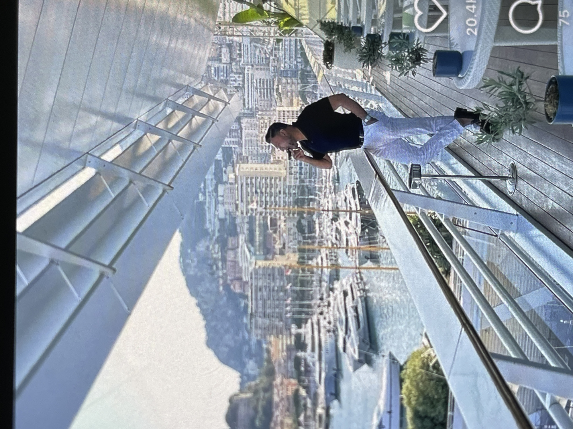 A man stands on a balcony, looking out at a city skyline and a harbor filled with yachts. He is wearing a black polo shirt, white pants, and black shoes. He is holding a cigar to his lips. The balcony has a wooden floor and a metal railing. There are plants in pots on the balcony, and a table and chairs. The city in the background is built on a hillside and is full of buildings of various sizes and colors. The harbor is filled with many boats, most of which appear to be yachts. The sky is clear and blue.