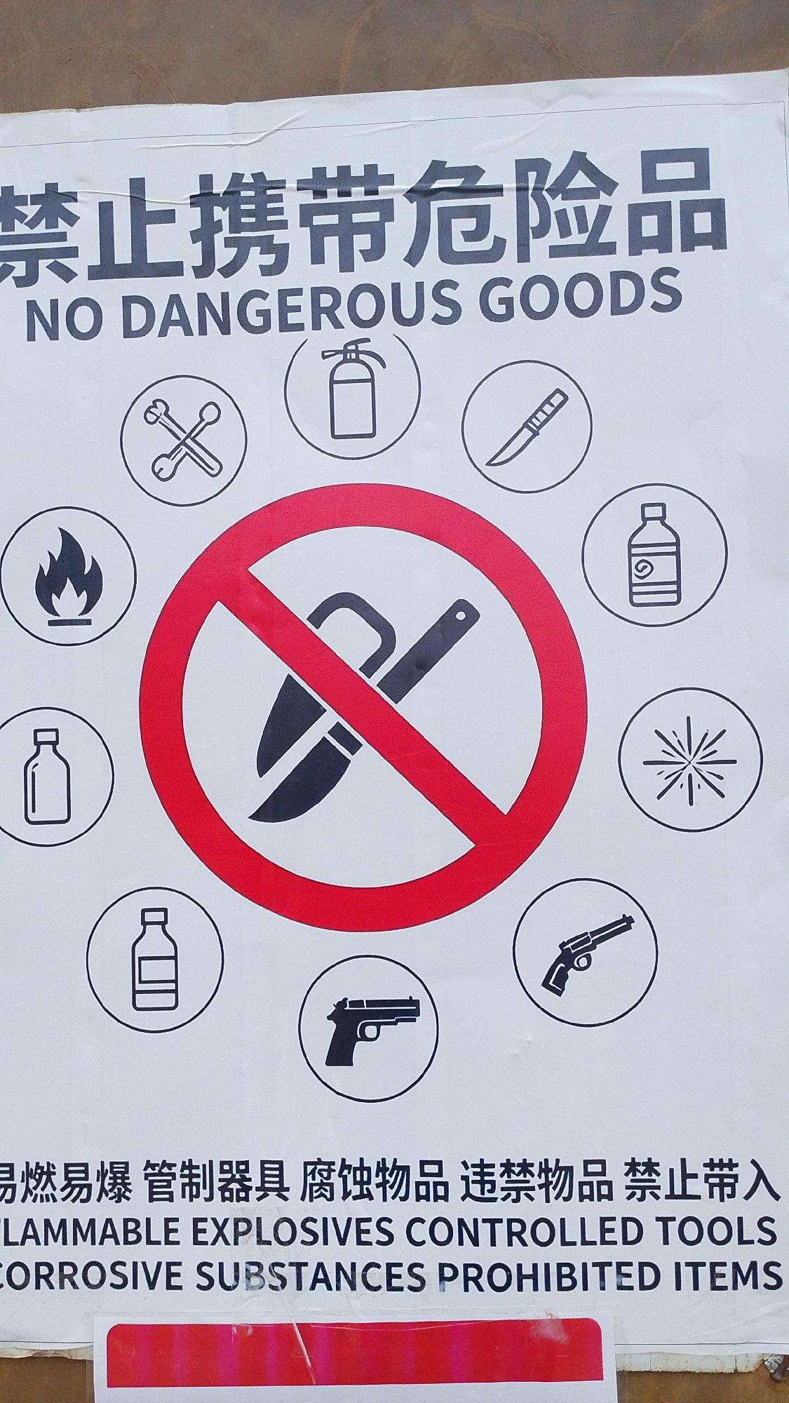 The image shows a sign with the text "NO DANGEROUS GOODS" in English, and Chinese characters above it. Below the English text are several circular icons depicting prohibited items, including a fire, a bottle, tools, a fire extinguisher, a knife, a bottle with a label, a spark, a handgun, and scissors. In the center of the sign, a larger red circle with a diagonal line across it contains an icon of scissors, indicating they are prohibited. The bottom of the sign also has text in English and Chinese listing categories of dangerous goods. The sign is mounted on a white surface, possibly a wall or a board. The background appears to be an outdoor environment with a textured surface, suggesting it is located in a public area. The overall scene depicts a safety warning sign.