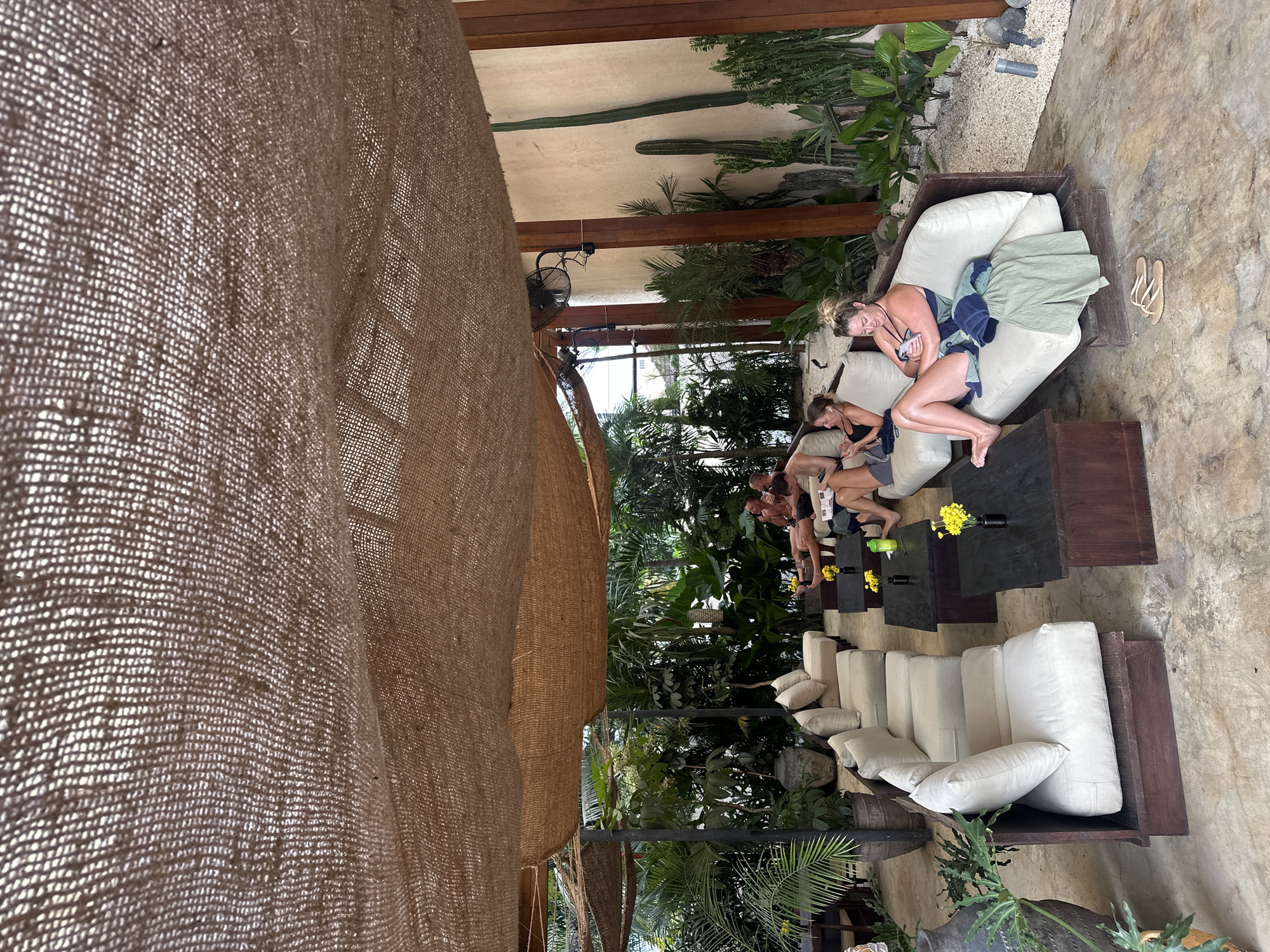 An outdoor seating area is covered by a woven, tan tarp. Several people are relaxing on low, cream-colored sofas arranged around dark wooden coffee tables. In the foreground, a woman in a blue swimsuit is sitting on a sofa, looking at her phone, with a towel draped over her legs. A pair of flip-flops rests on the concrete floor in front of her. Behind the sofas, lush green plants and several cacti are visible. The overall impression is a relaxed, tropical environment.
