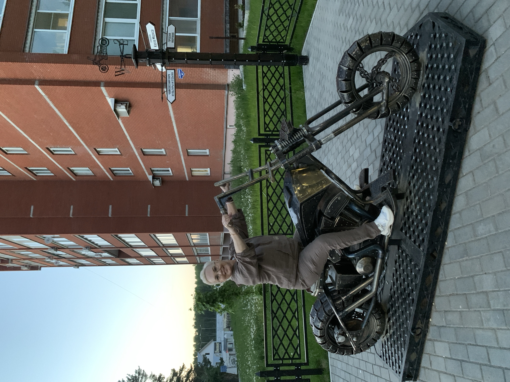 A woman is posing on a metal motorcycle sculpture in Chaikovsky, Russia. She is wearing brown clothes and white shoes, and is smiling at the camera. The motorcycle is black and appears to be made of recycled metal parts. The sculpture is displayed on a metal platform. In the background is a multi-story brick building, a street sign, a black metal fence, grass, and trees. The sky is light.