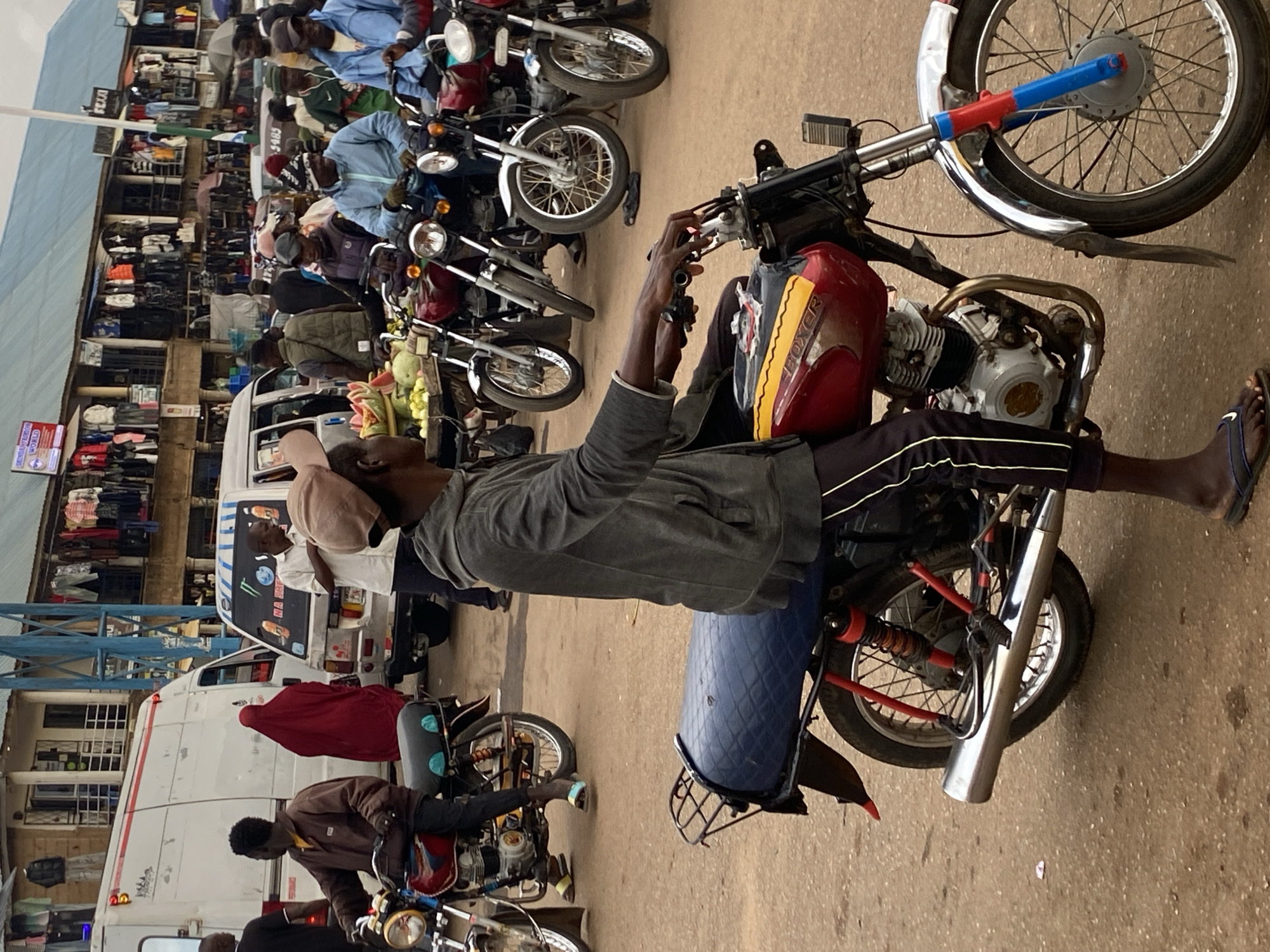 This image, set in Bukuru, Nigeria, captures a bustling street scene, seemingly a market or commercial hub, during daylight hours. The primary focus is on numerous motorcycles and the people associated with them, giving the impression of a busy transport or taxi stand.
The central subject is a man sitting on a red and black "BOXER" brand motorcycle, wearing a grey long-sleeved shirt, dark trousers, and flip-flops. His hands are on the handlebars. To his right, another motorcycle has a fruit stand attached, displaying watermelon and other green fruits. Many other motorcycles are visible, lined up or being ridden, predominantly red, black, or dark blue, often with chrome exhausts and spoke wheels. Several individuals, mostly men, are seen on or beside these motorcycles, dressed in casual wear including shirts, jackets, and caps. Some are wearing vibrant blue tops.
In the background, multi-story buildings line the street, featuring open-front shops displaying clothing and various goods. Blue structural elements are visible, likely part of awnings or building supports. White vans or minibuses are also present on the street, with one having "MONSTER" and "MA SHAA ALLAH" written on its side. The ground is a paved, reddish-brown surface.
Visible text includes "BOXER" on the fuel tank of the central motorcycle, "D734" and "5485" on signs attached to buildings in the background, and partial text "WORLD" and "EXPRESS" on a red and white sign. The overall atmosphere is one of active daily commerce and transport in an urban environment.