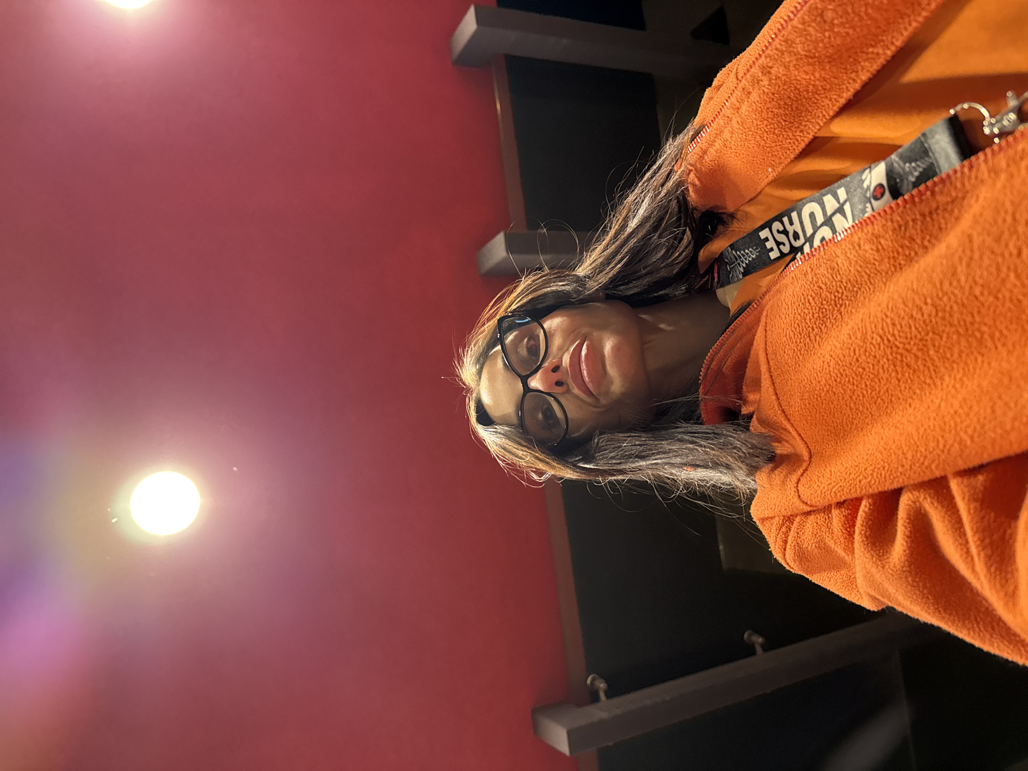 A woman wearing glasses and an orange fleece jacket with a "NURSE" lanyard is pictured from a low angle. She is positioned in front of a red wall with a light source shining on it. Adjacent to the red wall is a dark, possibly black, wall with wooden shelving or architectural details. While the city of Pleven, Bulgaria, is specified in the prompt, there is no visual evidence in the image to confirm the location.