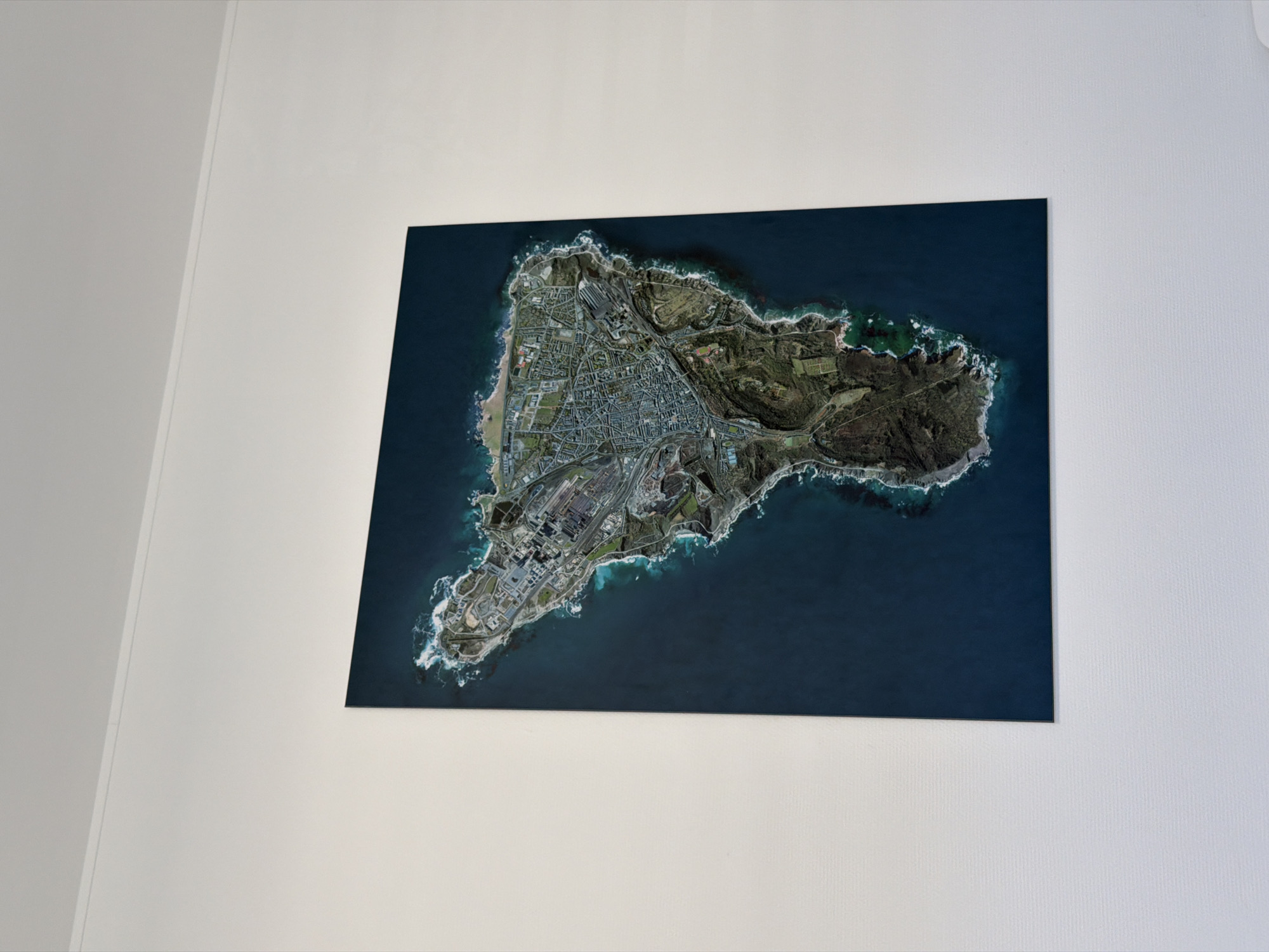 A framed aerial photograph of a peninsula hangs on a white wall. The peninsula features a mix of urban development, industrial areas, and natural landscapes. The city of Esch-sur-Alzette, Luxembourg, is not visible in the photograph.