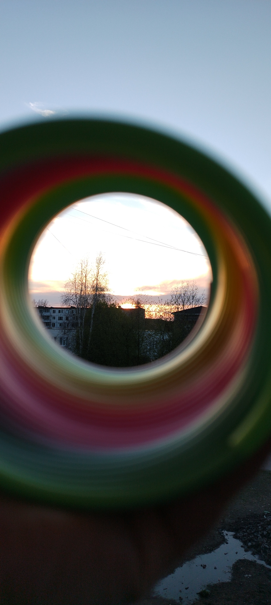 The image captures a landscape through a circular frame of an unidentified object. The frame consists of concentric rings of green, red, orange, and yellow. Seen through the frame is a low-rise building with multiple windows, and a few bare trees silhouetted against the sky. The sky displays the bright light of the setting or rising sun, with orange and yellow hues near the horizon. Above this is a blue sky with a thin strip of cloud. The foreground below the circular frame shows dark ground with a small puddle of water.