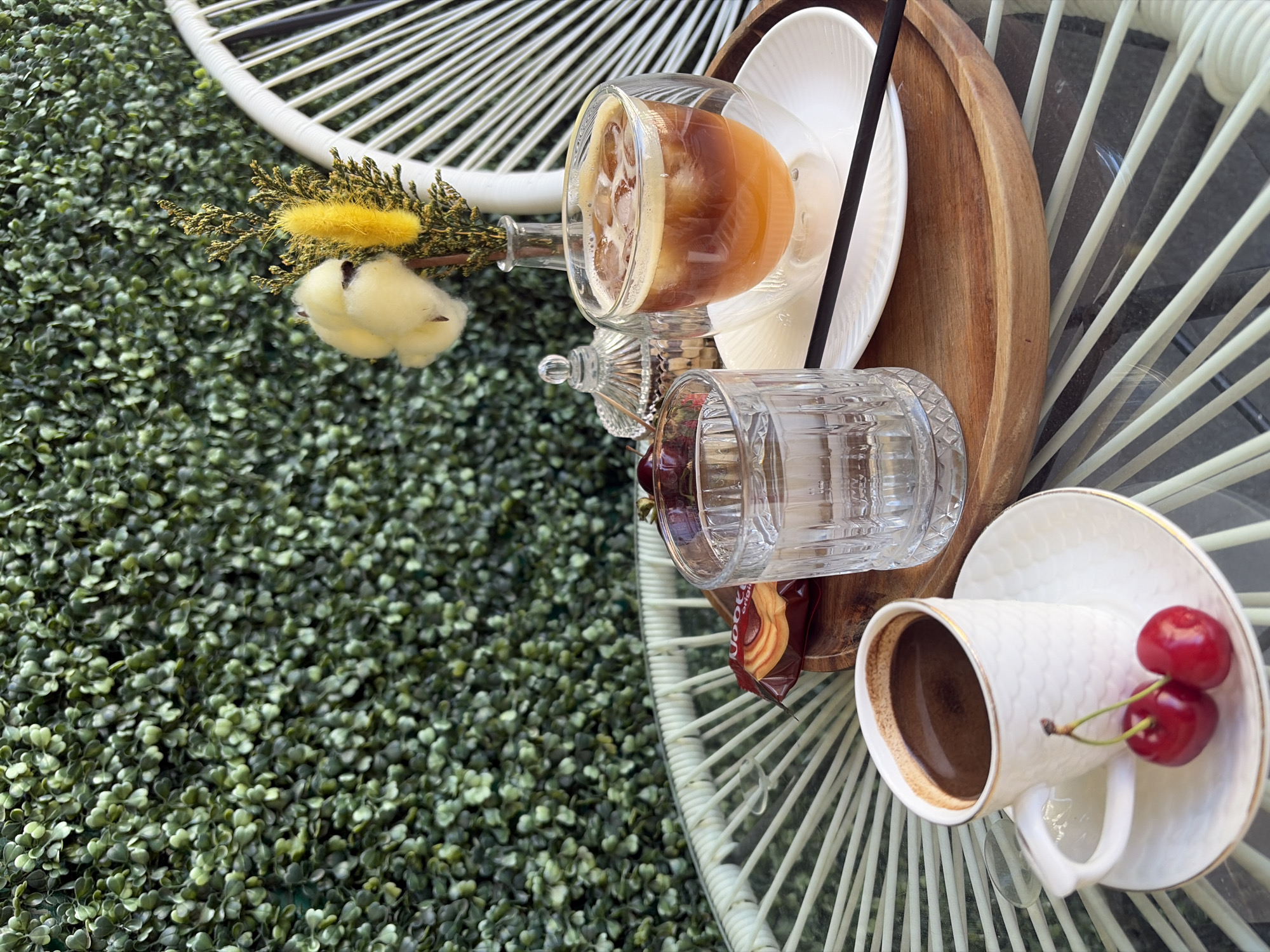 Captured from an elevated perspective, the scene presents an outdoor table arrangement, set against a backdrop of lush green foliage, possibly in Budva, Montenegro. On the table, a variety of refreshments are carefully arranged. A glass containing a light-colored beverage with ice sits alongside an empty, decorative glass. There's also a white cup of dark coffee placed on a saucer, adorned with two cherries. A wooden serving tray contains white plates and a straw. An unopened cookie package rests alongside the glasses. The table's glass top reflects its white, ornate metal frame, which, along with floral decorations, adds to the table's aesthetic appeal.