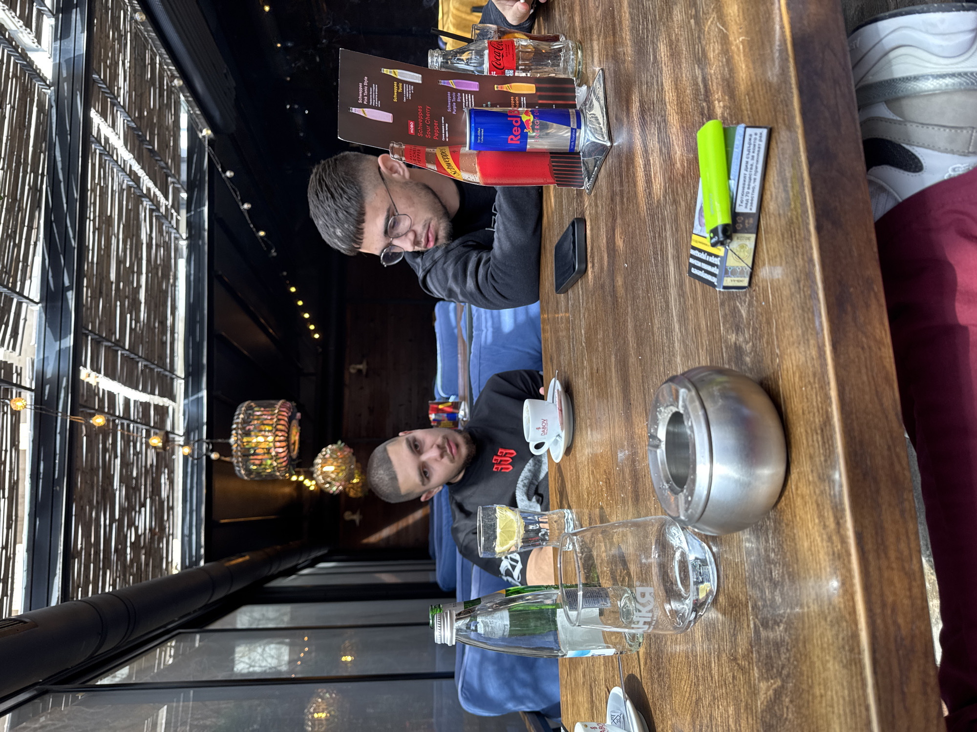 Here is a description of the image:
The image shows two men at a table in what appears to be a restaurant or cafe. One man is wearing glasses and a black shirt, looking at a beverage menu and holding a cell phone. The other man wears a black sweater with white symbols and seems to be drinking coffee.
On the table, there are various objects including beverage containers (Red Bull, Schweppes, Coca-Cola), water bottles, glasses, a lighter, a pack of cigarettes, an ashtray, a coffee cup, and a menu stand. The table itself is wooden.
The cafe appears to have an outdoor or semi-outdoor setting, with views of the outside and unique decorative lighting. The cafe seems to be located in Pleven, Bulgaria, based on the Cyrillic text on the cigarette pack. Also, the word DABOV on the coffee cup is a well known coffee brand from Bulgaria.