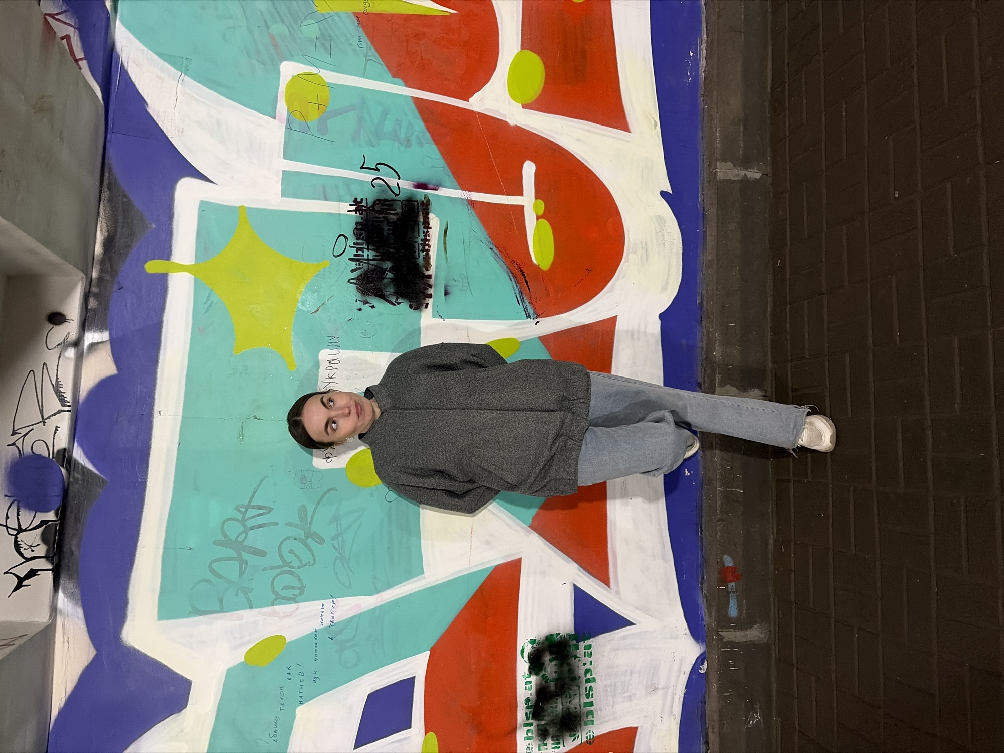 A young woman in light blue jeans, white sneakers, and a dark grey jacket stands in front of a large, colorful graffiti mural in Izhevsk, Russia. The mural has large geometric shapes in shades of blue, white, red, green, and yellow. There is also some black tagging and other writing on the mural in what appears to be a Cyrillic alphabet. The woman is centered in the image, with her head and torso in front of the mural and her lower legs extending to the right, where a dark brick or stone wall meets the edge of the graffiti. The lighting is somewhat dim.