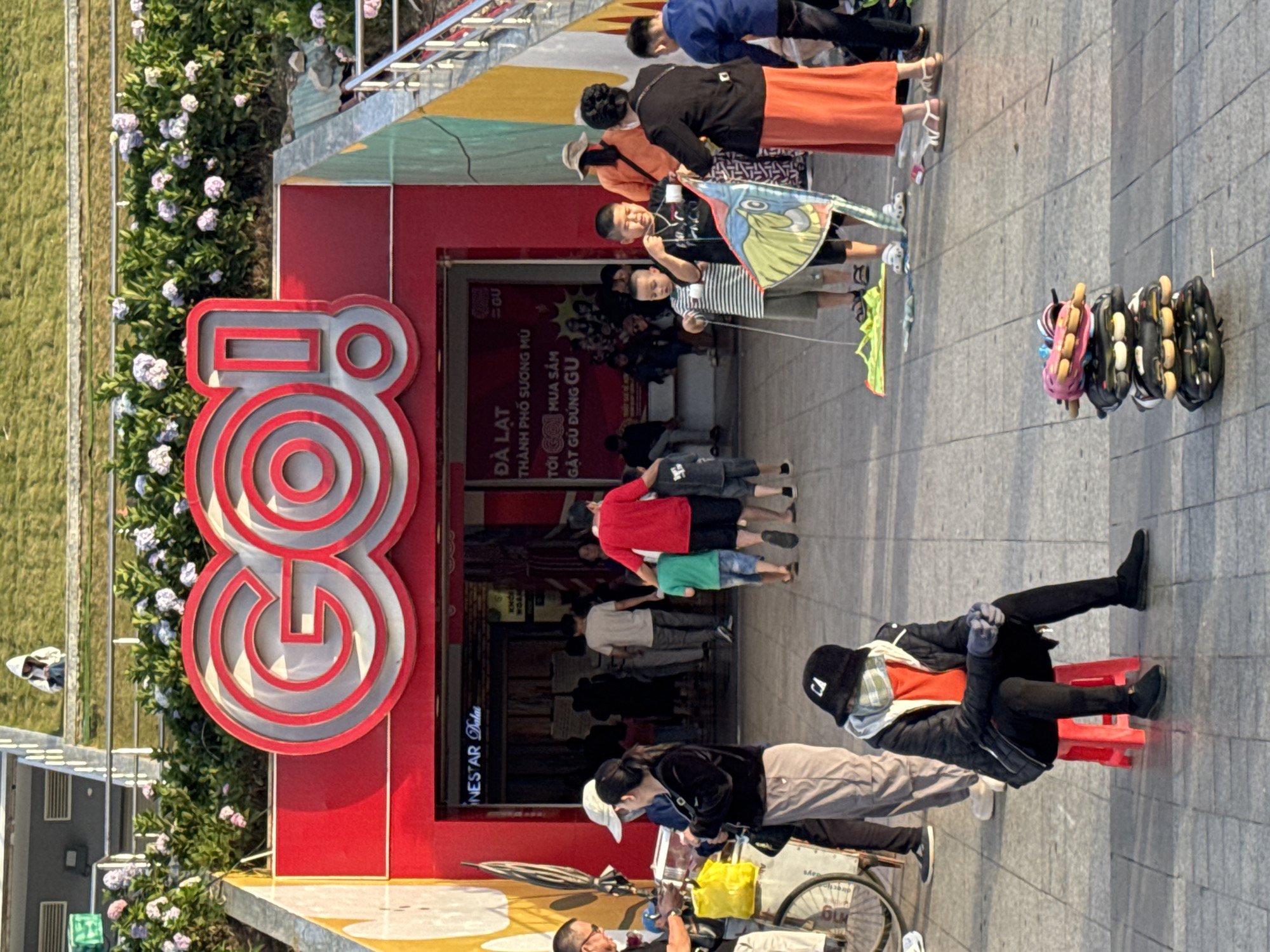 The image depicts a street scene in front of a store with a large red "GO!" sign. The store appears to be a commercial establishment located in Da Lat, Vietnam, as suggested by text on a poster within the store's entrance. A group of people are gathered outside the store. In the foreground, a person wearing a black jacket, orange shirt, and a face mask sits on a red stool. To the right of the seated person, a stack of roller skates is displayed on the ground. Further into the scene, several children are interacting, with one child holding a colorful kite. Adults are also present, some observing the children, while others are engaged in conversation or shopping. The background shows the store's entrance and a grassy area above. The environment appears to be an urban outdoor space, possibly a pedestrian area, with a paved ground surface.