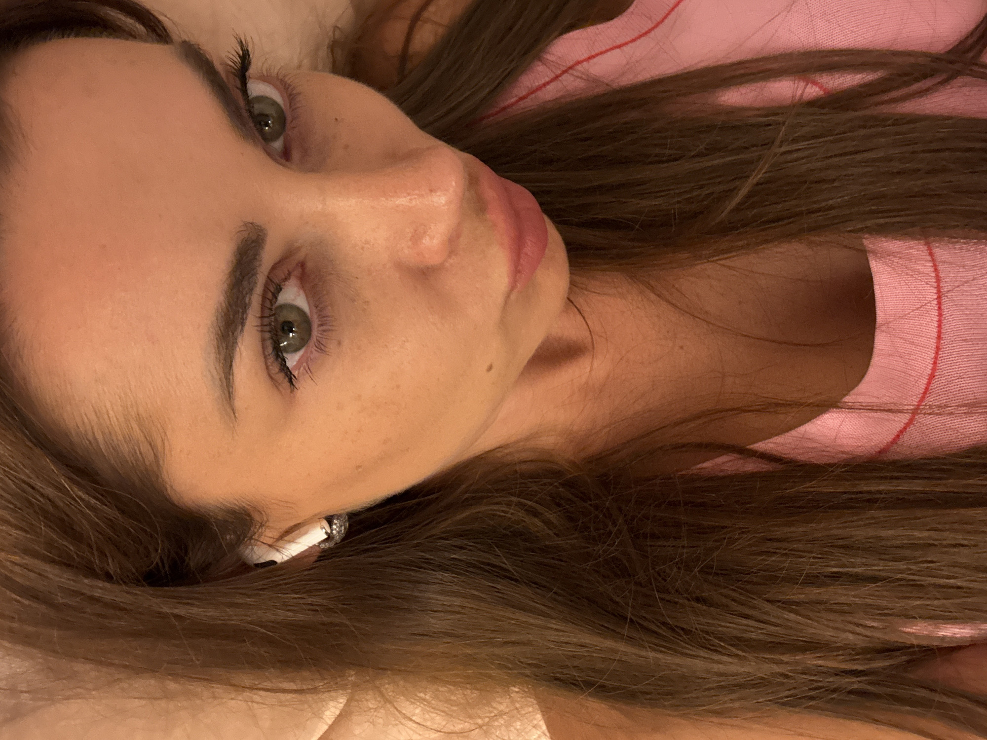 A close-up, high-angle selfie of a woman with long, dark brown hair, green eyes, and fair skin. She is wearing a pink top and white wireless earbuds. The woman's head is tilted, and her gaze is directed downwards. The lighting appears to be artificial, casting a warm glow on her skin. The background is indistinct and appears to be a textured surface, possibly bedding or upholstery. There is no discernible location in Rome, Italy.