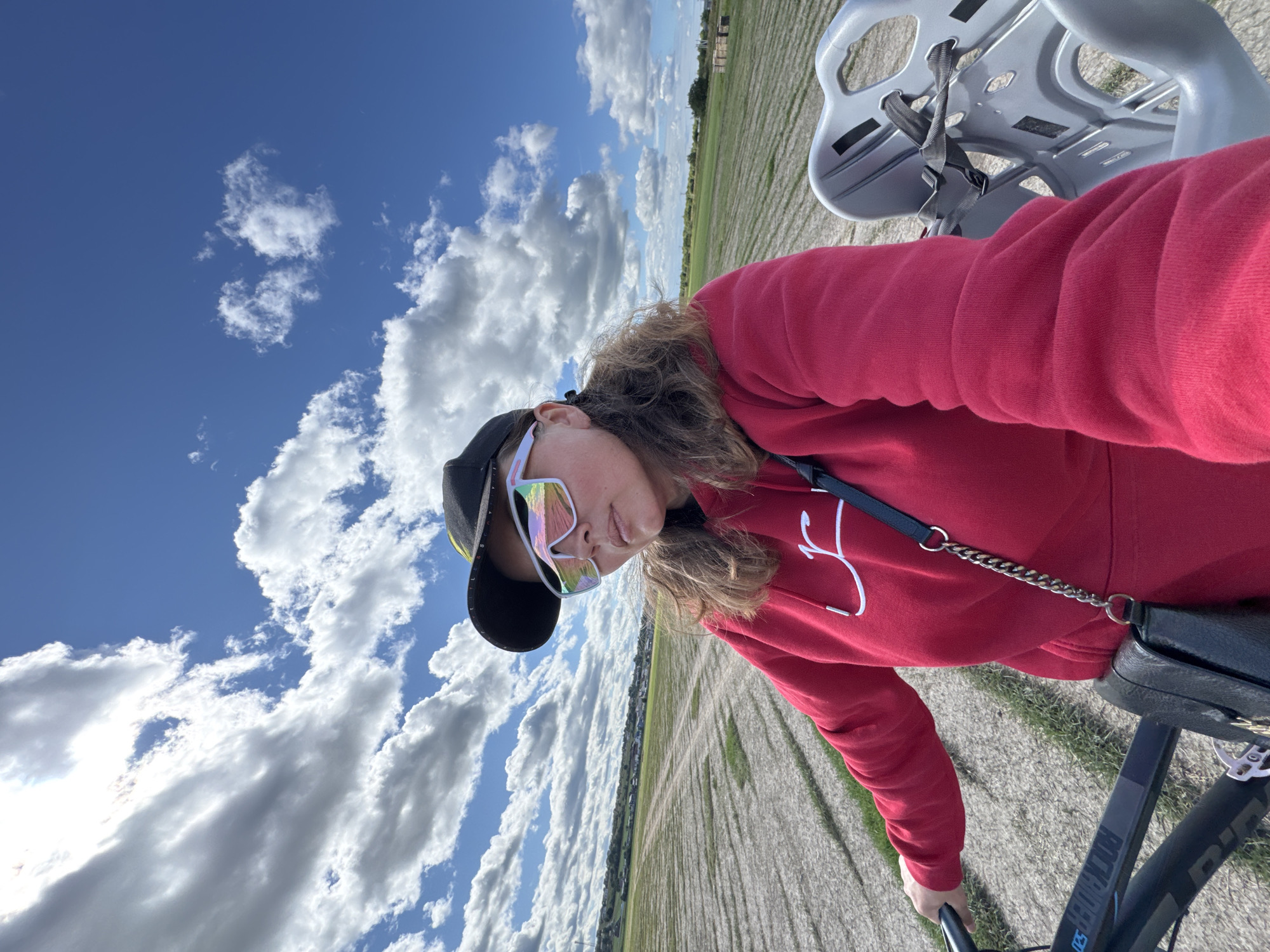 A woman wearing a red hoodie, a black baseball cap, and reflective sunglasses is on a bicycle in an open field under a cloudy blue sky. The field appears to be cultivated with visible rows. In the background, there is a line of trees and buildings. A child's bicycle seat is attached to the rear of the woman's bicycle. The image is a selfie, indicating the woman is taking the photo herself. The location is identified as Kempelevo, Russia.