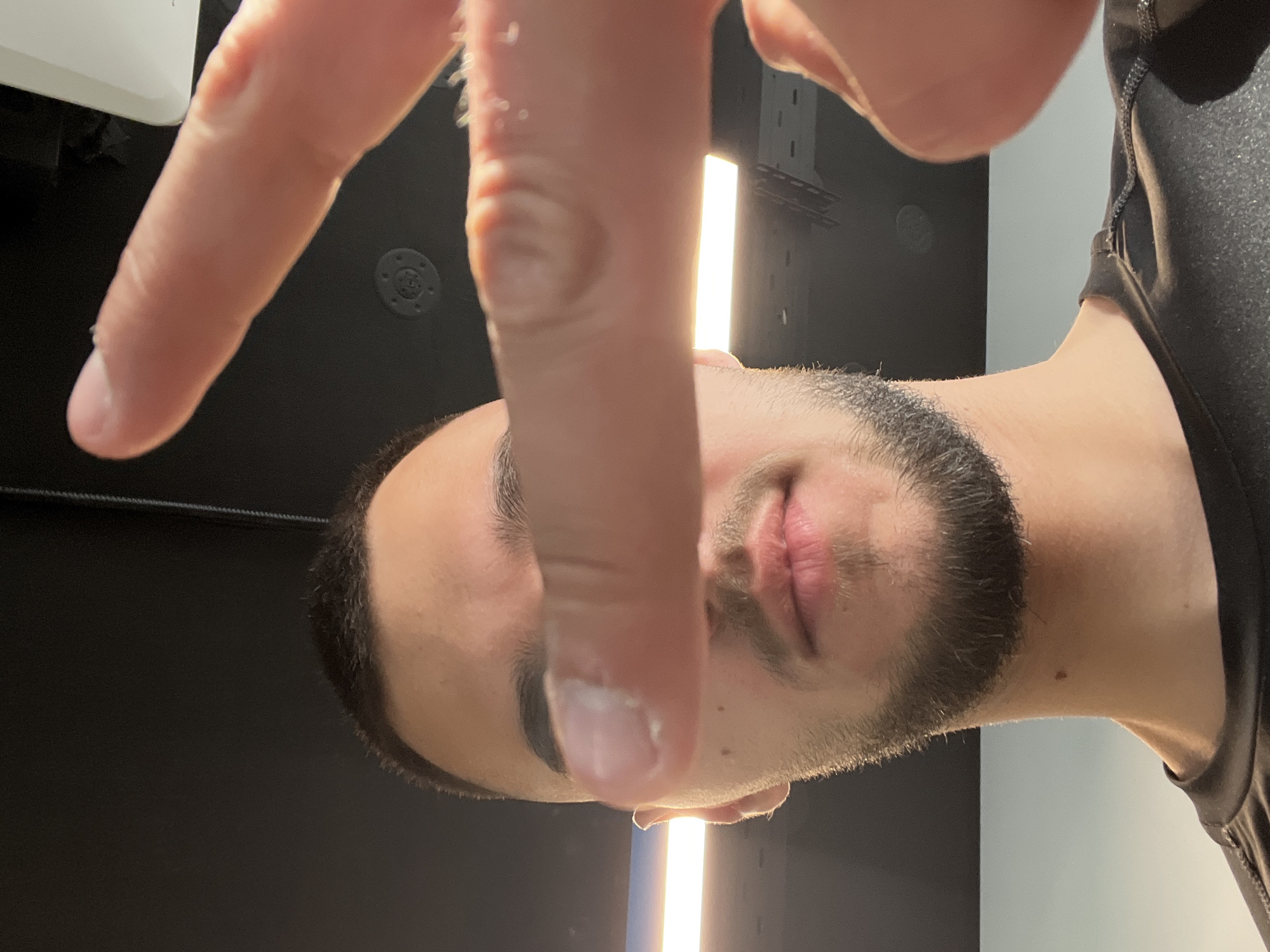 A close-up, chest-up shot captures a man indoors, looking towards the camera. His right hand, with the index finger and thumb prominently featured in the foreground, partially obscures his face. He has a neatly trimmed dark beard and mustache. His lips are slightly parted, revealing no particular expression. He is wearing a dark, possibly athletic, short-sleeved top with visible stitching details.
The background consists of a dark wall or surface. A bright, vertical strip light illuminates the space behind his head and right shoulder. In the upper left portion of the background, a white, rectangular object is visible, possibly a light fixture or equipment. Another dark, circular object with a patterned surface is visible on the wall behind his hand. The skin on his fingers shows some texture, appearing slightly dry or flaky in places. The overall lighting suggests an artificial, indoor environment, possibly a studio or a modern interior. No text is visible in the image.