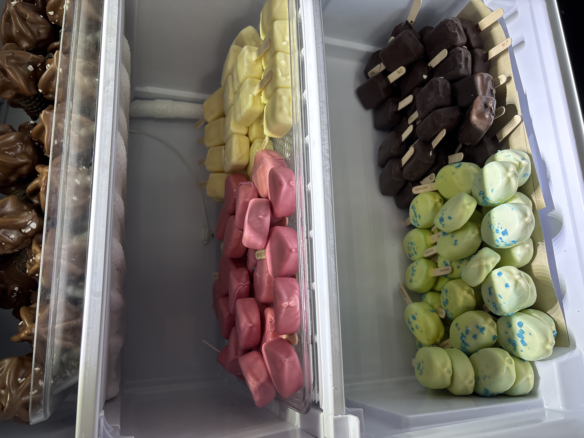 The image shows three freezer compartments filled with different types of popsicles. The left compartment contains what appear to be chocolate-covered pretzels with sea salt. The middle compartment holds yellow square popsicles on top, followed by pink square popsicles. The right compartment contains dark chocolate square popsicles above light green round popsicles with blue spots. The popsicles are arranged in plastic containers. The scene is likely a shop or market selling frozen treats, location is unknown, but the product labels are in Greek, not Bulgarian.