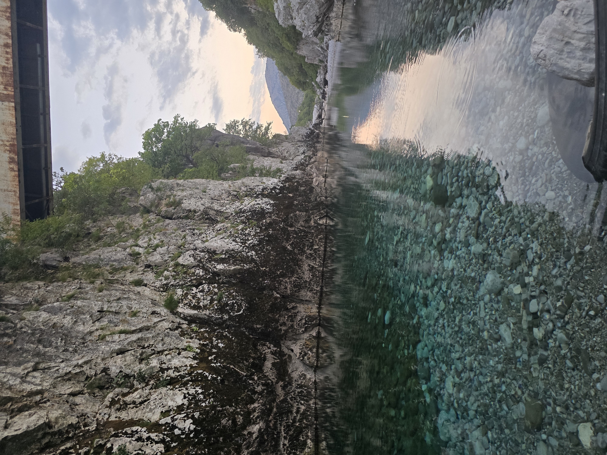 The image depicts a scenic river canyon in Bioče, Montenegro. A clear, shallow river flows through the canyon, with its rocky bottom visible through the transparent water. Steep, moss-covered rock faces rise on either side of the river, with trees and vegetation clinging to their upper edges. A bridge, possibly made of concrete and steel, spans the top of the canyon, with its structure partially visible. The sky above is overcast with some patches of sunlight breaking through, casting a soft glow on the water and surrounding landscape. The overall impression is one of natural beauty and tranquility.