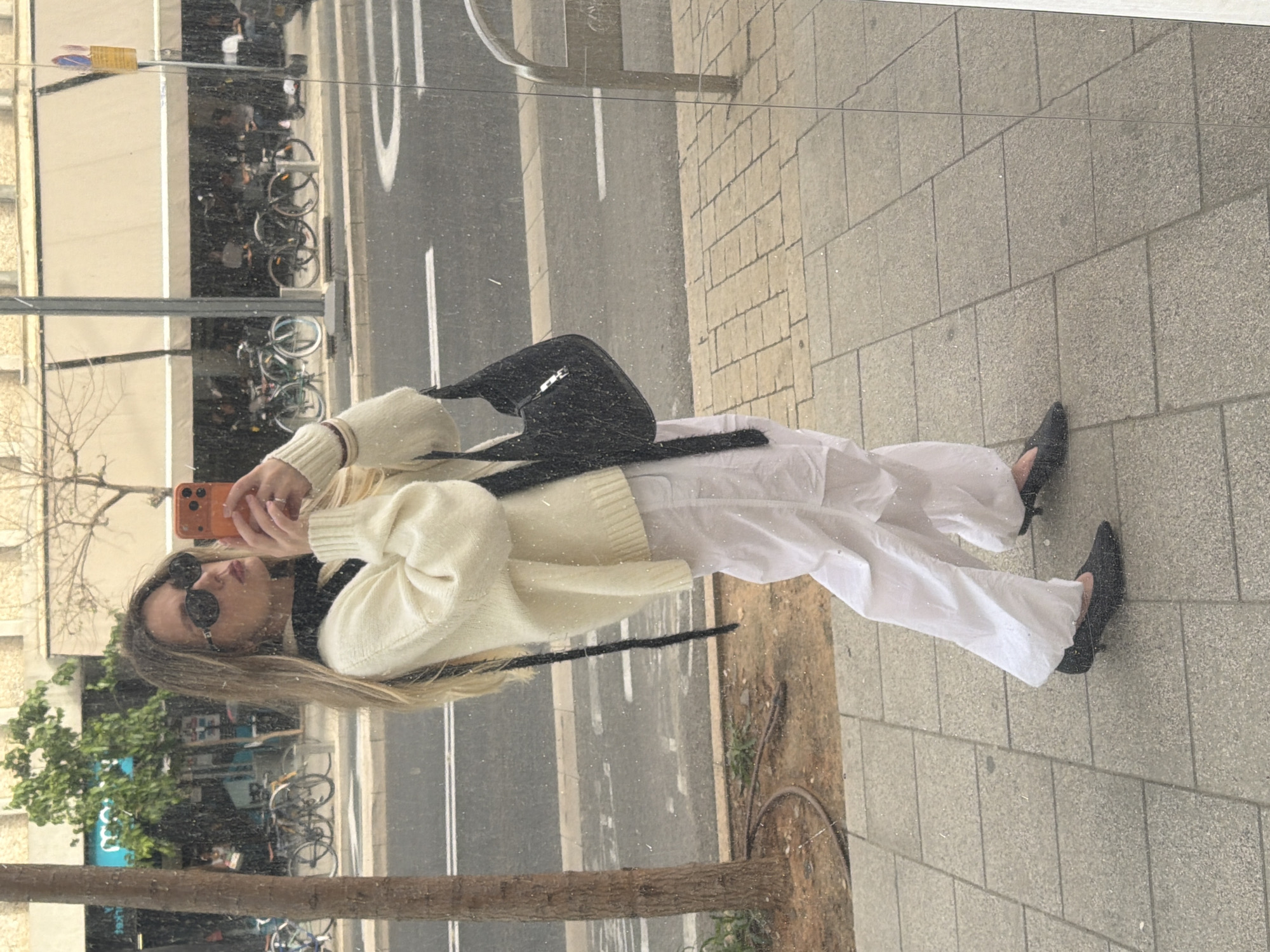 A woman is reflected in a glass pane, possibly a shop window, on a street in Tel-Aviv, Israel. The reflection includes the street, bicycles parked nearby, and trees. The woman is holding a cell phone as if taking a picture. She is wearing sunglasses, a light-colored sweater, white pants, and black pointed-toe shoes. She has a black purse over her shoulder. The sidewalk is made of gray paving stones. The street behind her has white lane markings. A segment of a tree trunk is visible at the bottom of the image, indicating its position relative to the glass pane.