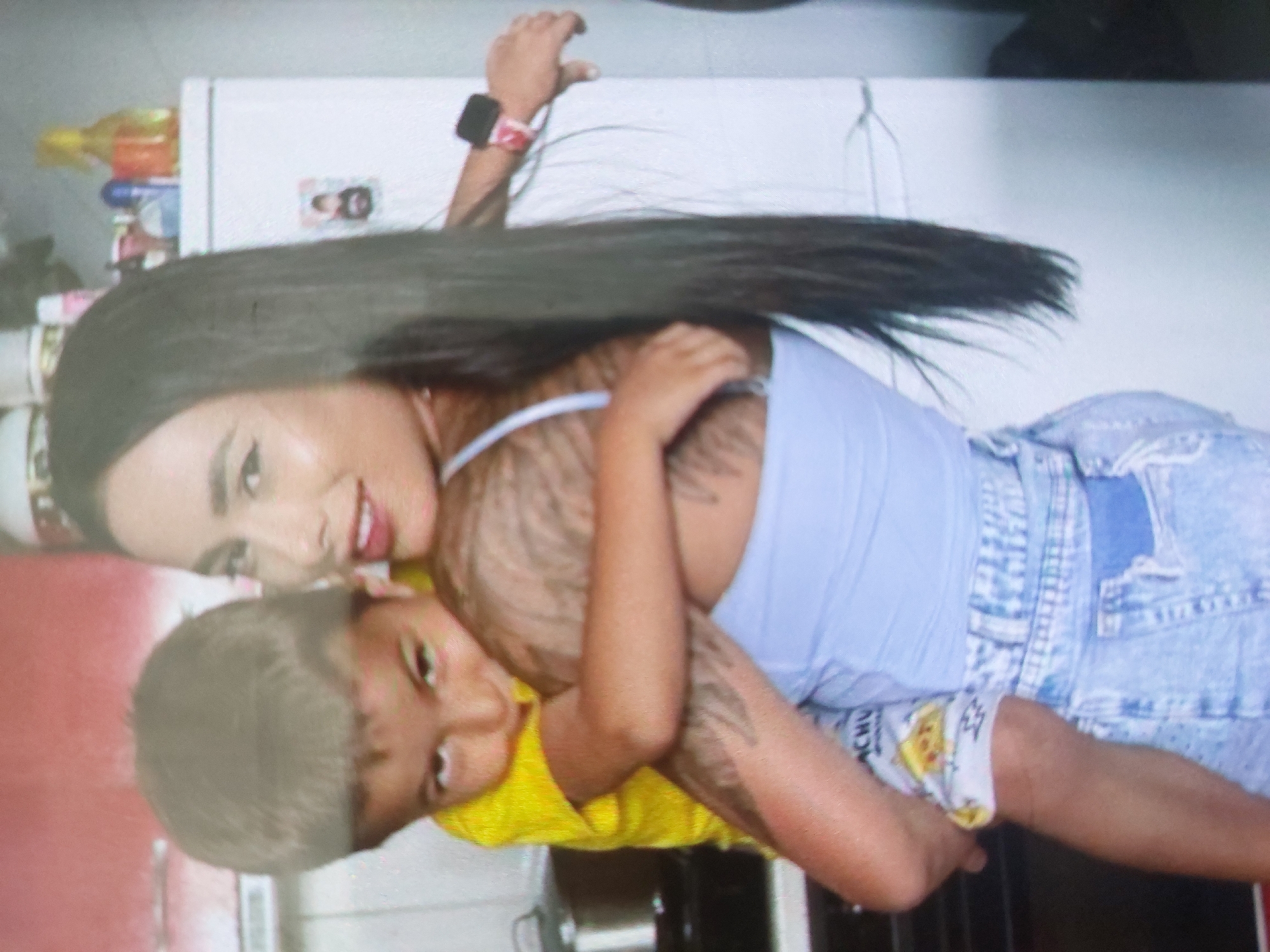 The image depicts a woman with long, dark hair, wearing a light blue tank top and denim shorts, holding a young child with light gray hair who is wearing a yellow shirt. The background includes a white refrigerator and various items on top of it. The location, Tsarevo, Bulgaria, cannot be verified from the image.