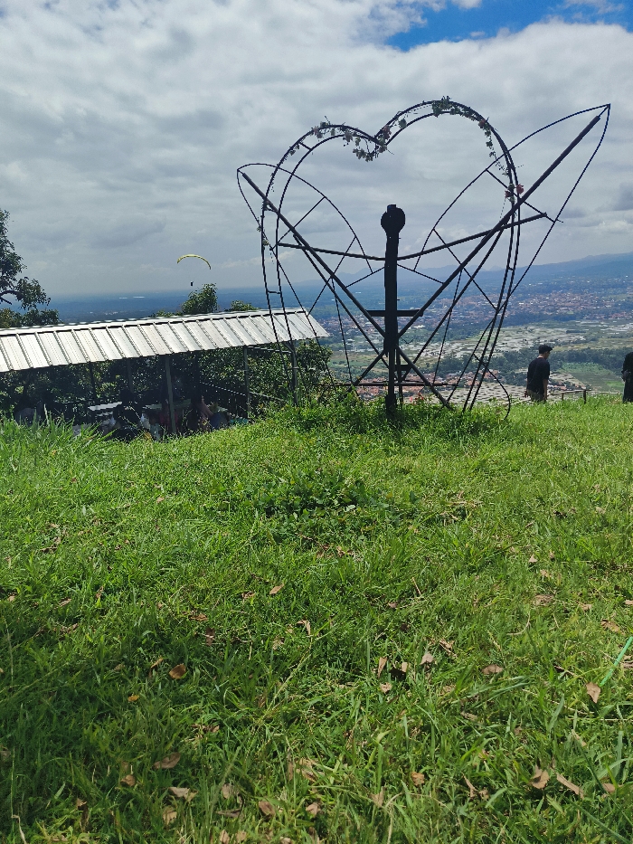 The image shows a grassy hilltop overlooking a city in Majalengka, Indonesia (latitude: -6.846, longitude: 108.204). There is a large metal heart-shaped structure with a thick metal pole in the center. A small white pavilion-like structure sits in the foreground, with a few people inside, some sitting on benches and others standing. A single paraglider appears as a small dot in the distance. Two people are visible in the foreground, standing in front of the metal structure. One person is facing the camera, and the other has their back turned to the camera. The hilltop is covered in lush, green grass with scattered brown leaves. The sky is blue with scattered white clouds. The image was taken on a sunny day.