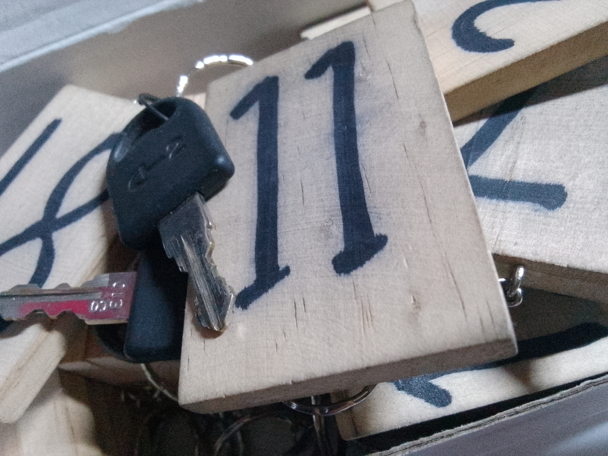 In a close-up, low-angle shot, a wooden key tag with the number "11" spray-painted in dark blue is prominently displayed, slightly off-center to the right. The tag is part of a larger collection of similar wooden tags, some of which are visible in the background, also bearing painted markings. A black plastic key fob is attached to the tag via a silver ring. Partially obscured by the tag and fob, a silver key with a red painted section and some indiscernible numbers or markings is visible. The objects are situated within a shallow, cardboard-like container, suggested by the visible edges and textures of the material. The lighting appears to be overhead, casting subtle shadows and highlighting the textures of the wood and metal. The scene is static and offers no information about human presence or activity in the city of Trenggalek, Indonesia.