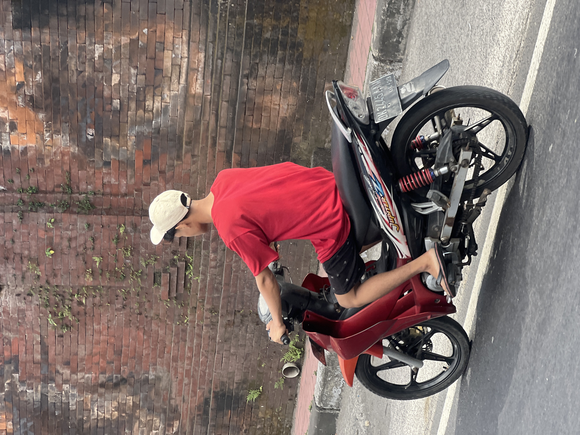 A young male wearing a red t-shirt and black shorts rides a red and black motorcycle on a paved road in Denpasar, Indonesia. He wears a beige baseball cap. The motorcycle has a license plate that reads "DK 2445 AN" and "06-23". The background features a weathered red brick wall with some small green plants growing on it. The road has a white line marking the lane. A concrete curb separates the road from a sidewalk. The overall scene depicts a common urban street view.