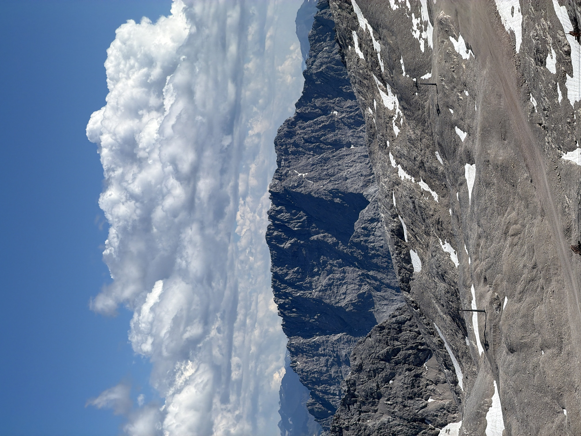 The image shows a mountainous landscape with a rocky, barren terrain dotted with patches of snow. In the foreground, the ground is covered in gravel and small rocks, with several areas of snow. A dark, tall, slender pole-like structure stands on a mound in the middle ground. Further back, a formidable mountain range with jagged peaks dominates the scene. The sky above is a vibrant blue, filled with large, fluffy white clouds. The overall impression is one of a high-altitude, alpine environment.
This scene is likely located in the vicinity of the Zugspitze, Germany's highest peak, which is a prominent landmark near Garmisch-Partenkirchen. The presence of ski lifts or similar infrastructure, hinted at by the pole in the middle ground, further supports this location, as the area is a popular ski resort. The rugged terrain and the presence of snow even during what appears to be a clear, sunny day are characteristic of the alpine environment found in this region. There are no people visible in the image.