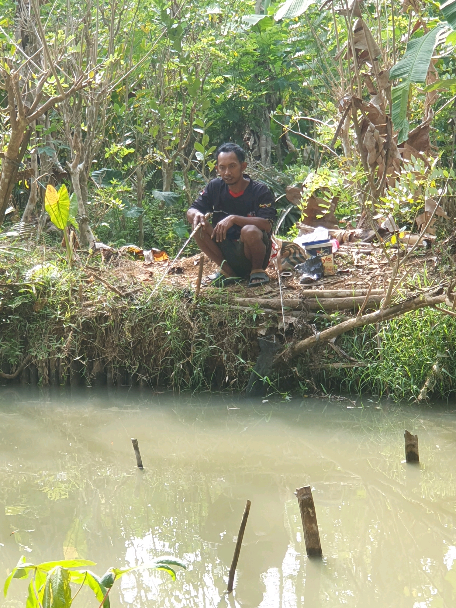 A man is sitting by a muddy canal in Pucungkerep, Indonesia, fishing with a rod. He is wearing a black t-shirt with red trim and dark shorts. He is crouching on the bank, surrounded by dense foliage and trees. A white bucket with the brand name "Dulux" is visible on the bank near him, along with other debris. The water is murky and reflects the surrounding greenery. Several wooden stakes are submerged in the canal.