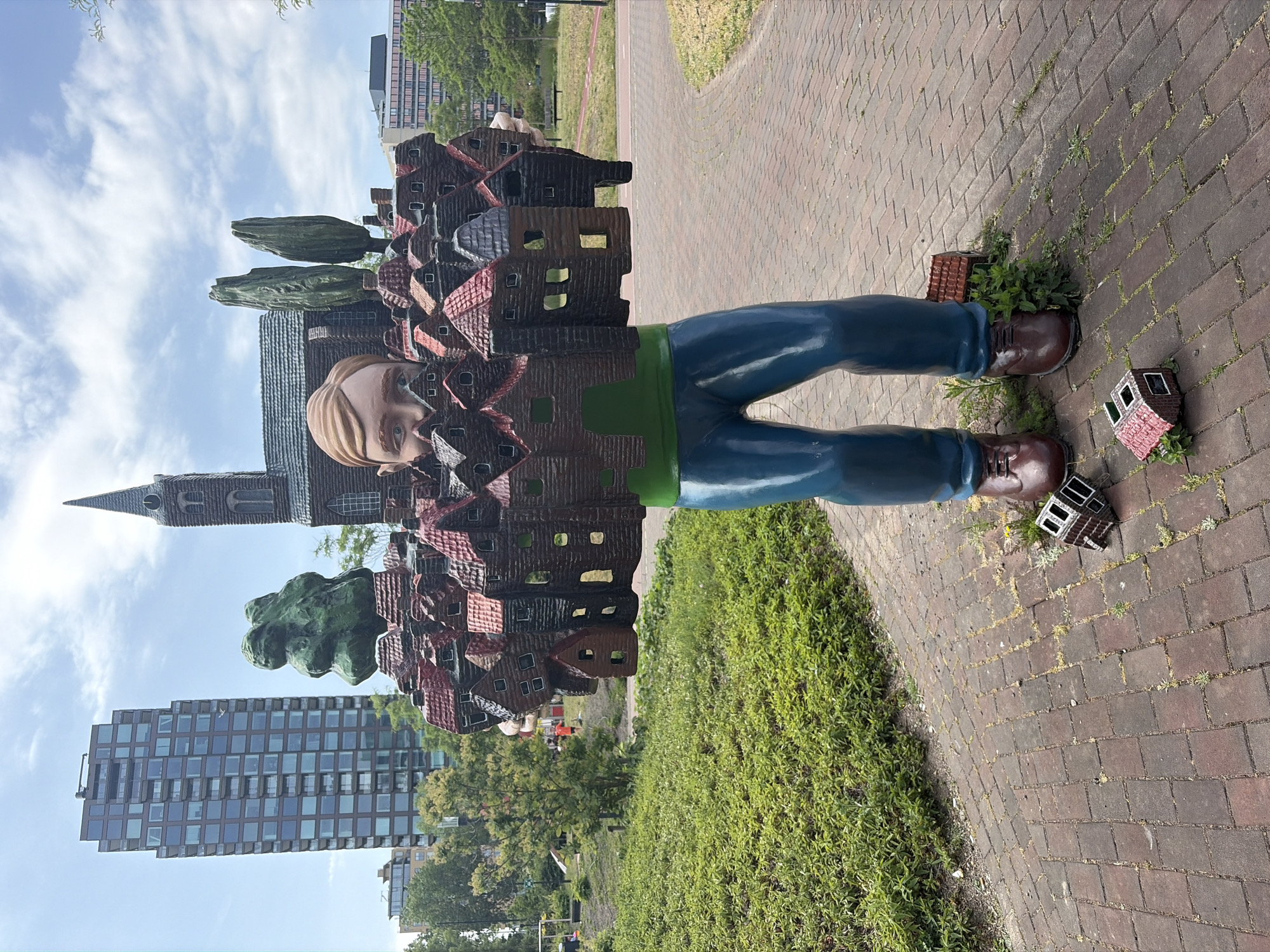 The image shows a statue of a man whose body is composed of houses and buildings, situated in Rijswijk, Netherlands. The statue has a head, blue legs, and brown shoes. Some of the houses composing his body have red roofs. A few detached house figures lie on the red brick path by his feet. Grass grows around the statue and through cracks in the brick path. A modern building and a church tower are visible in the background. The sky is partly cloudy.