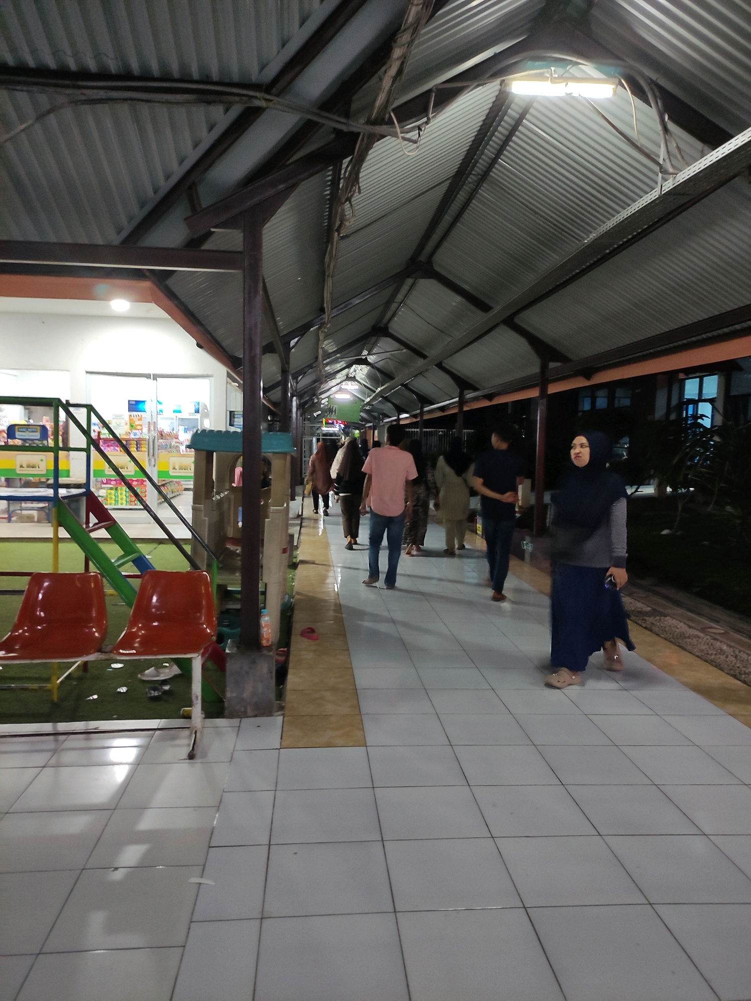 The image captures a covered walkway or corridor in Bulukumba, Indonesia, likely at dusk or night, given the artificial lighting and dark background. The walkway features a corrugated metal roof supported by dark metal beams, with fluorescent lights illuminating the path. On the left, a children's playground structure with colorful slides and a small playhouse is visible, adjacent to two empty red plastic chairs. The walkway itself is paved with light-colored square tiles, with a border of darker tiles. Several individuals are walking along the covered path. In the foreground on the right, a woman wearing a dark hijab and a long blue skirt is walking away from the camera. Further down the walkway, other people are seen walking in both directions. The environment appears to be a public space, possibly near a recreational area or a building with shops.