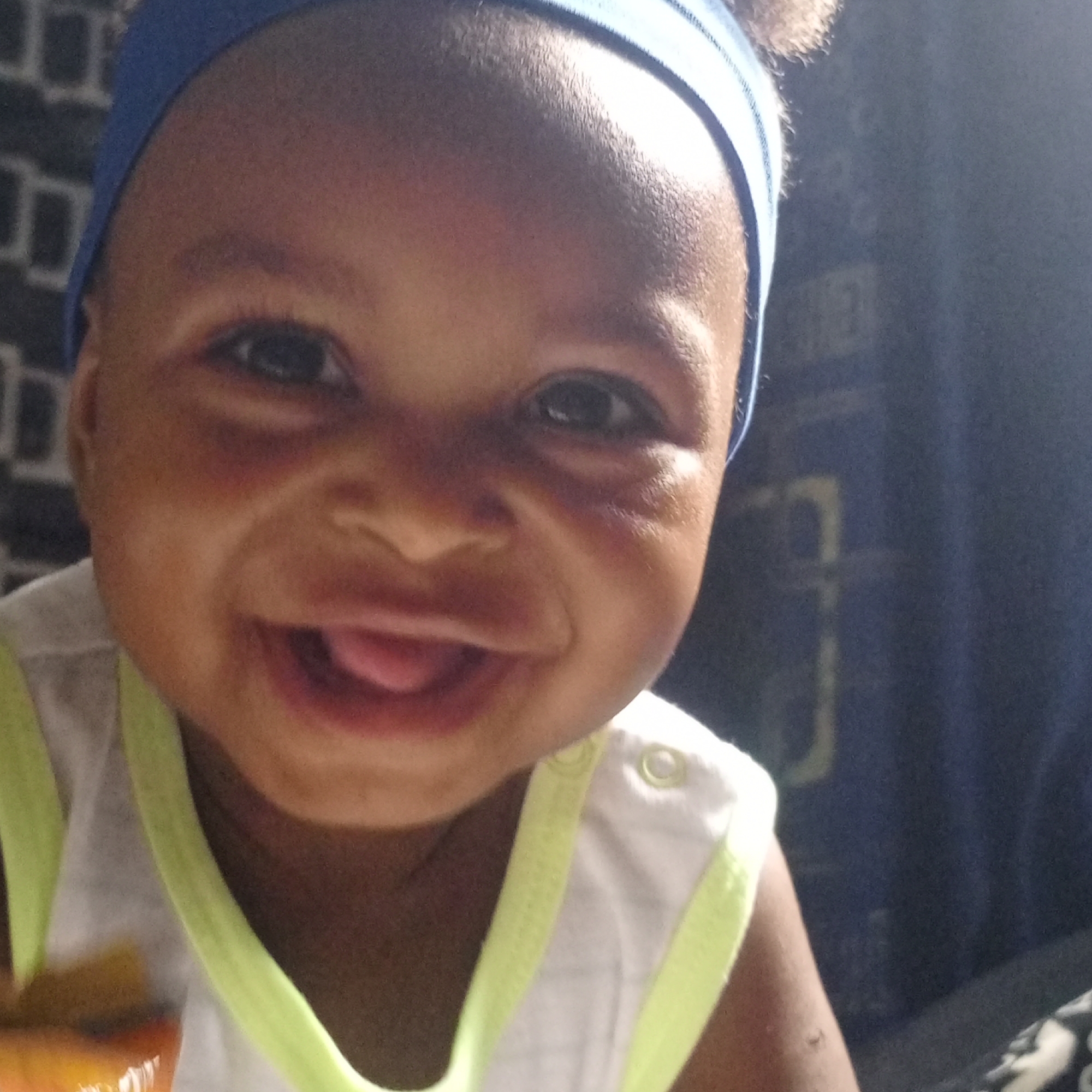 A close-up portrait captures a joyful baby or toddler, looking directly at the camera with a wide, open-mouthed smile, revealing their tongue. The child has dark skin and is wearing a blue fabric headband. Their eyes are dark and expressive, reflecting happiness. They are dressed in a white sleeveless top with bright neon yellow trim around the neckline and armholes, with a snap closure visible on the shoulder. A blurry, orange-yellow object, possibly food, is partially visible in the bottom left corner of the frame. The background is dark and out of focus, with faint patterns suggesting a patterned wall or curtain indoors. The lighting is soft and even, highlighting the child's cheerful expression. No specific time of day or external weather can be determined from the indoor setting. There is no visible text.