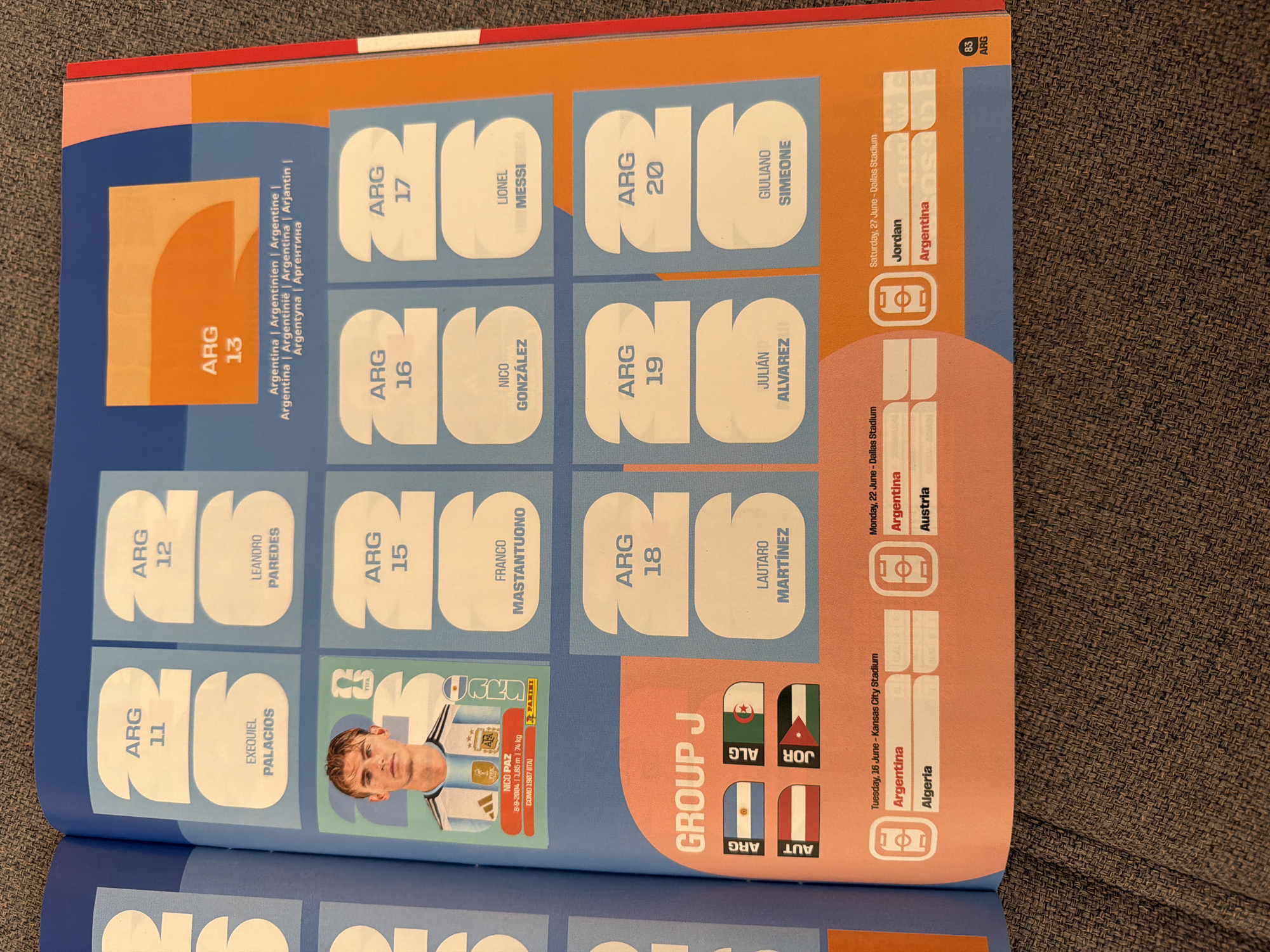 The image is a close-up, high-angle shot of an open page from a sticker album, likely depicting a soccer team or tournament. The page is primarily light blue and orange, with white cutouts for stickers. The left side of the page shows a player sticker of Nico Paz, an Argentinean player, with his name and biographical details. Above his sticker are two large white sticker slots labeled "ARG 11" and "ARG 12", with spaces for "EXEQUEL PALACIOS" and "LEANDRO PAREDES" respectively.
The right side of the page features larger white sticker slots, also with the "ARG" prefix, labeled "ARG 13", "ARG 15", "ARG 16", "ARG 17", "ARG 18", "ARG 19", and "ARG 20". Some of these slots have player names filled in: "FRANCO MASTANTUONO", "NICO GONZALEZ", "LIONEL MESSI", "LAUTARO MARTINEZ", "JULIÁN ALVAREZ", and "GIULIANO SIMEONE". The "ARG 13" slot has an orange background with text indicating "Argentina | Argentinien | Argentine | Argentina | Argentinie | Argentina | Arjantin | Argentyna | Аргентина".
At the bottom left of the page, under the heading "GROUP J", are four small flag stickers: Argentina, Algeria, Austria, and Jordan. Below these flags are schedule details for matches involving these countries.
The page is resting on a textured, dark gray surface that appears to be fabric, possibly a couch or a blanket. The lighting suggests an indoor environment.
There is no information in the image that connects it to Zlatitrap, Bulgaria.