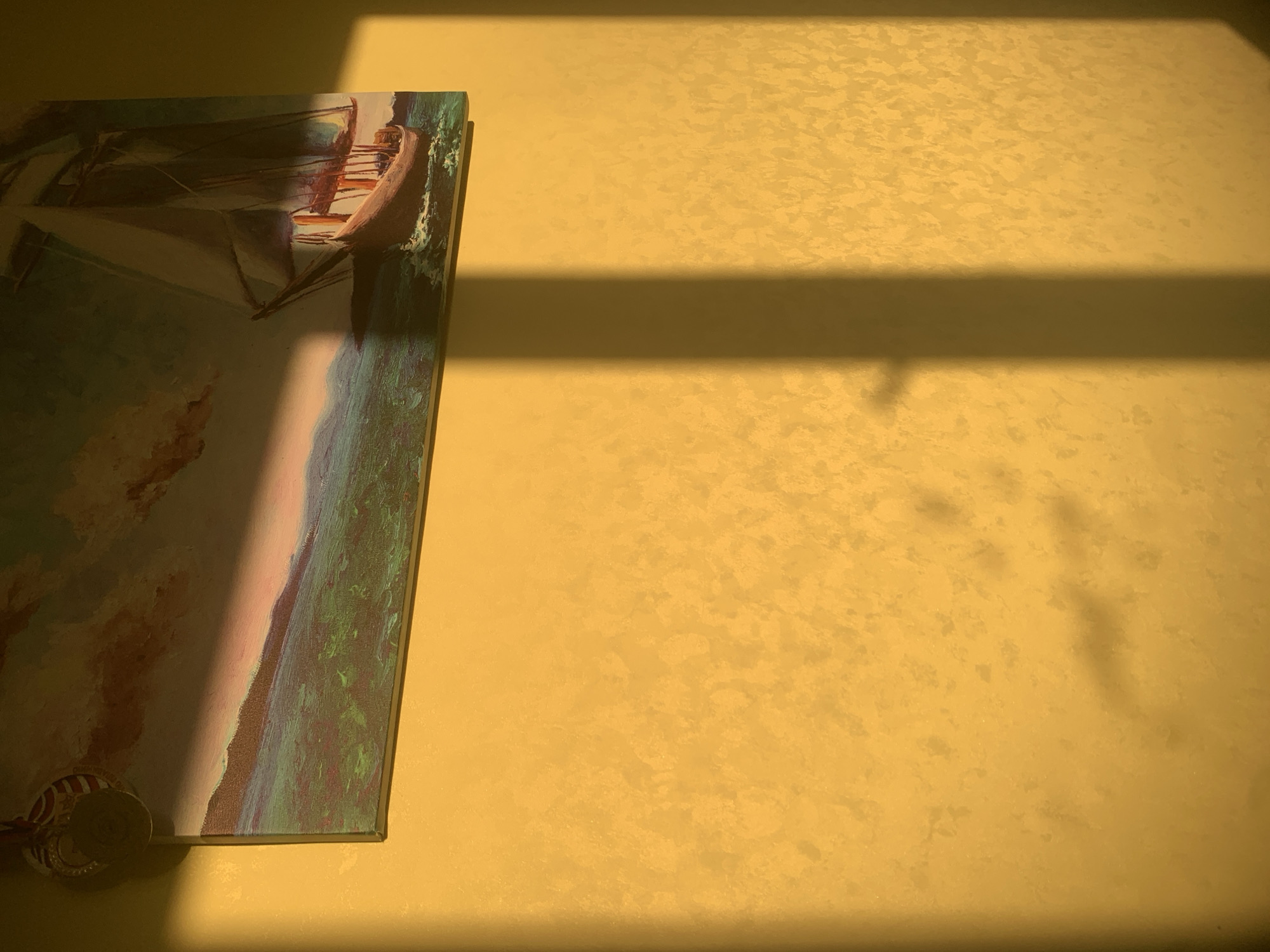 The image shows a close-up of a wall with a painting of a sailboat partially visible on the left side. The wall is textured and painted in a yellow hue. A dark shadow crosses the wall horizontally, created by an unseen object. At the bottom left corner of the painting, there are two coins. The location is impossible to determine as the image provides no information about the location.